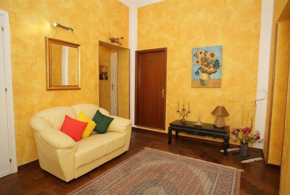 A room with a beige sofa, a wooden door, a painting of sunflowers, and a small table with a lamp and flowers. - Hotel Florio (Palermo)