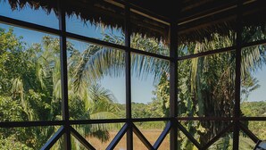 Una terraza de madera con una mesa y dos sillones de descanso, con vista a un paisaje tropical con palmeras y un río.