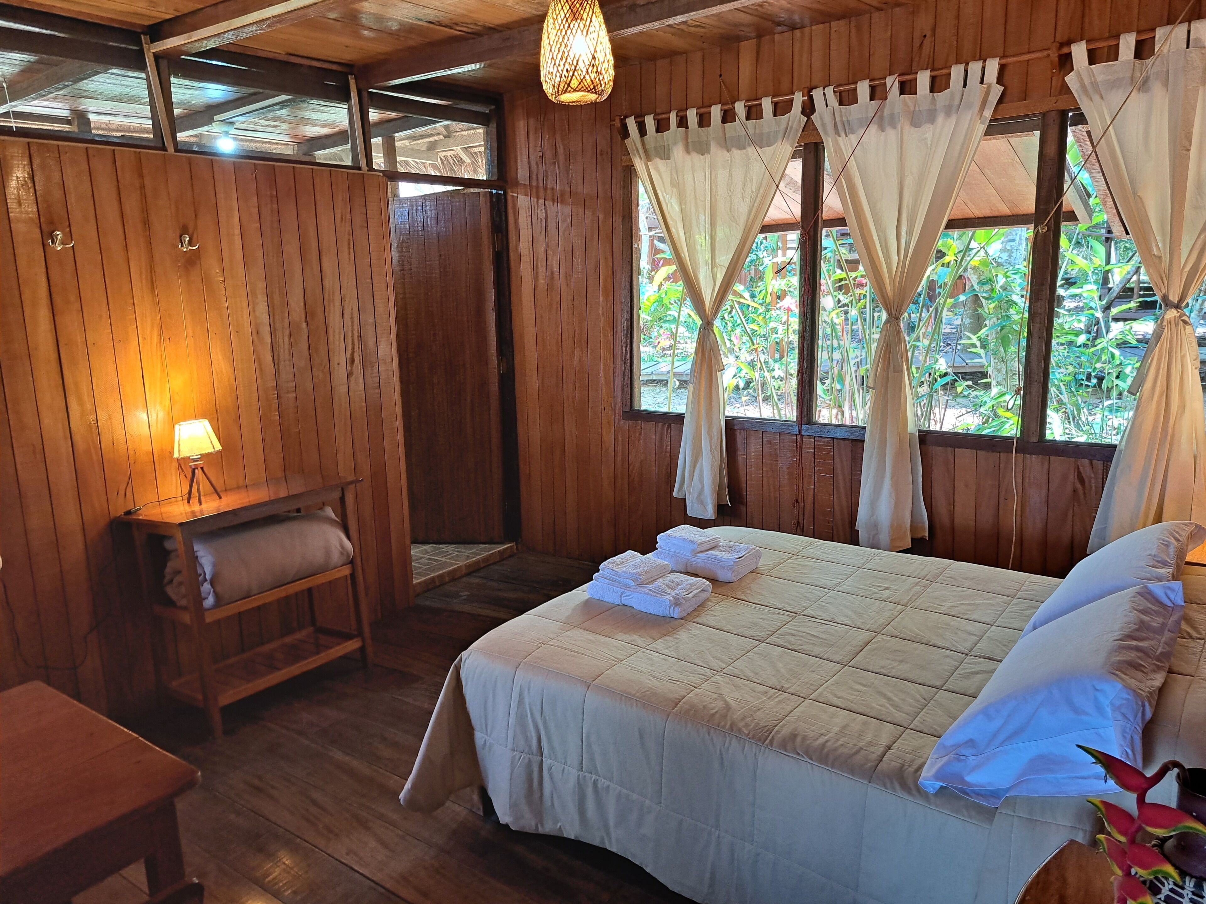 Una habitación de madera con una cama, un banco, una mesa y una lámpara.