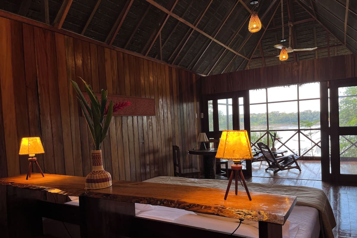 A room with wooden walls, a large bed, two bedside lamps, a small table, a chair, and a view of the outdoors.