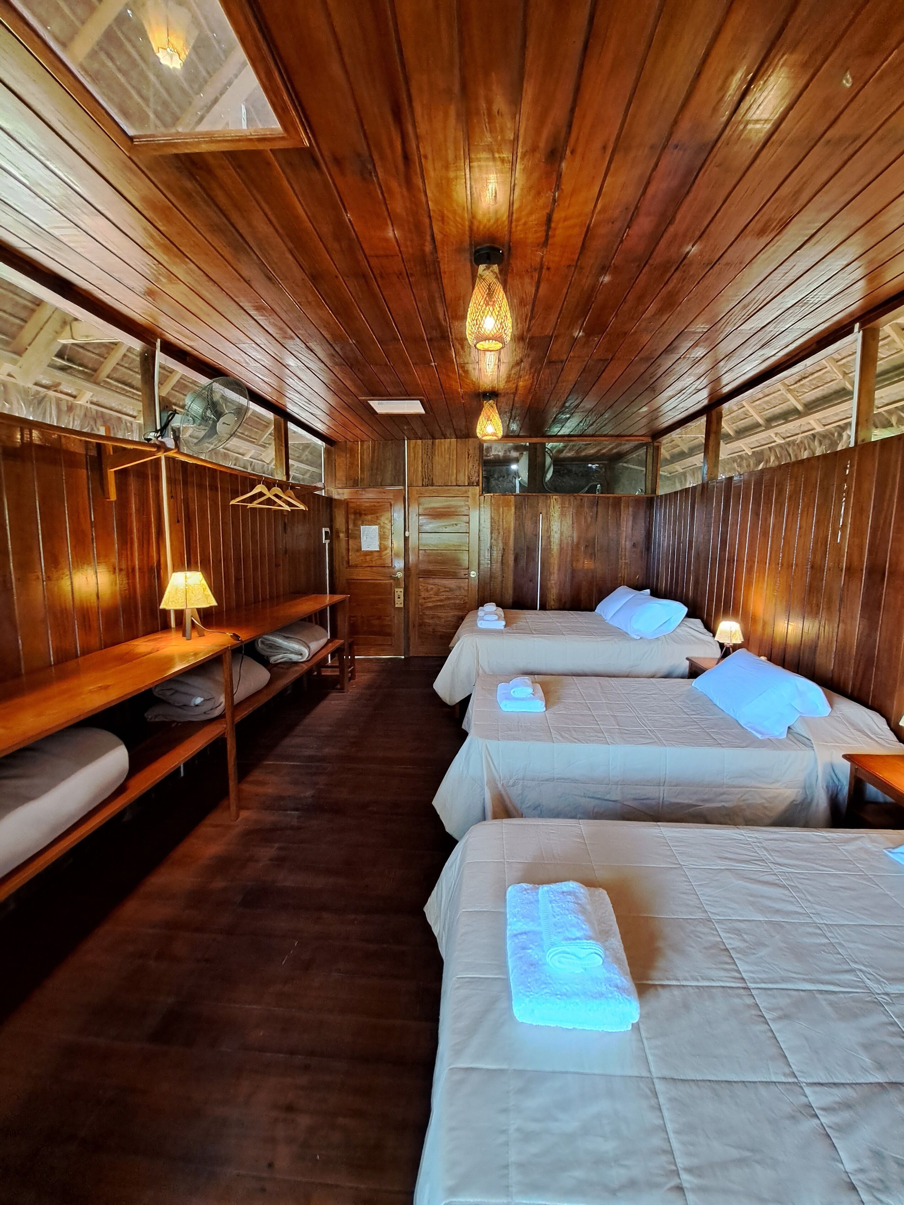 A wooden cabin with four beds, a bench, a fan, and a table with a lamp.