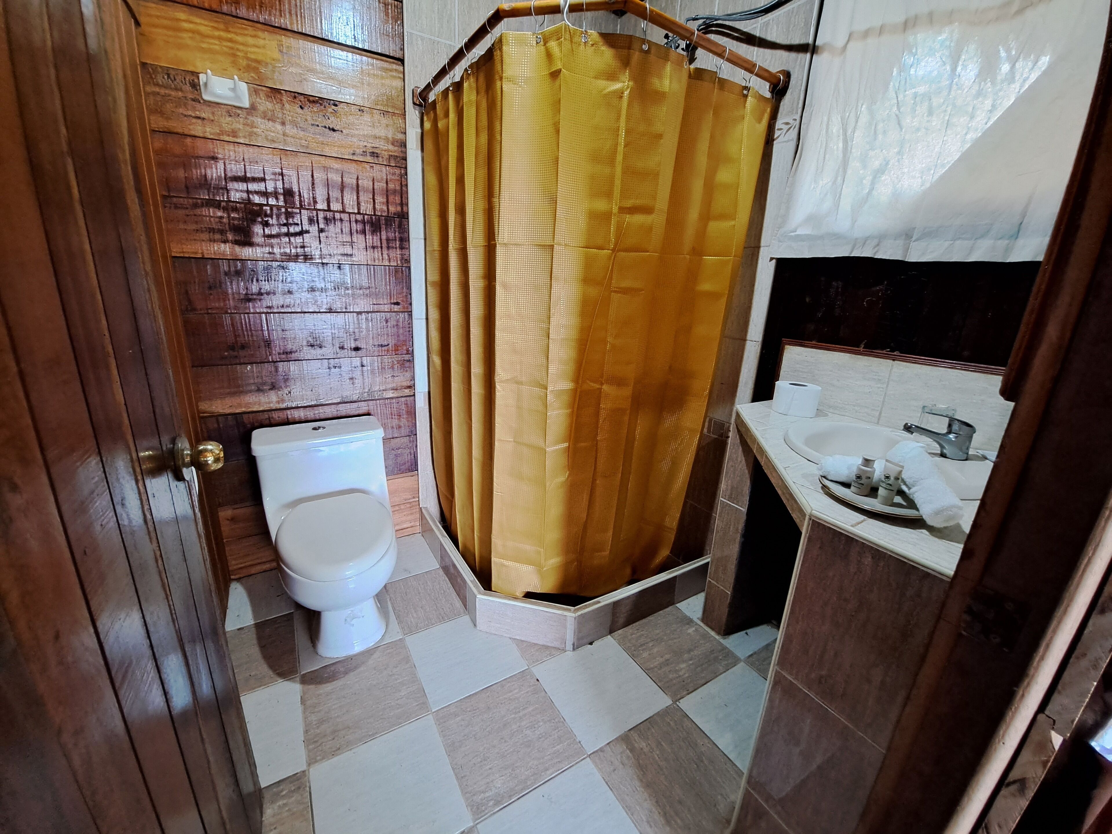 A bathroom with a wooden wall, a white toilet, a yellow shower curtain, and a sink with toiletries.