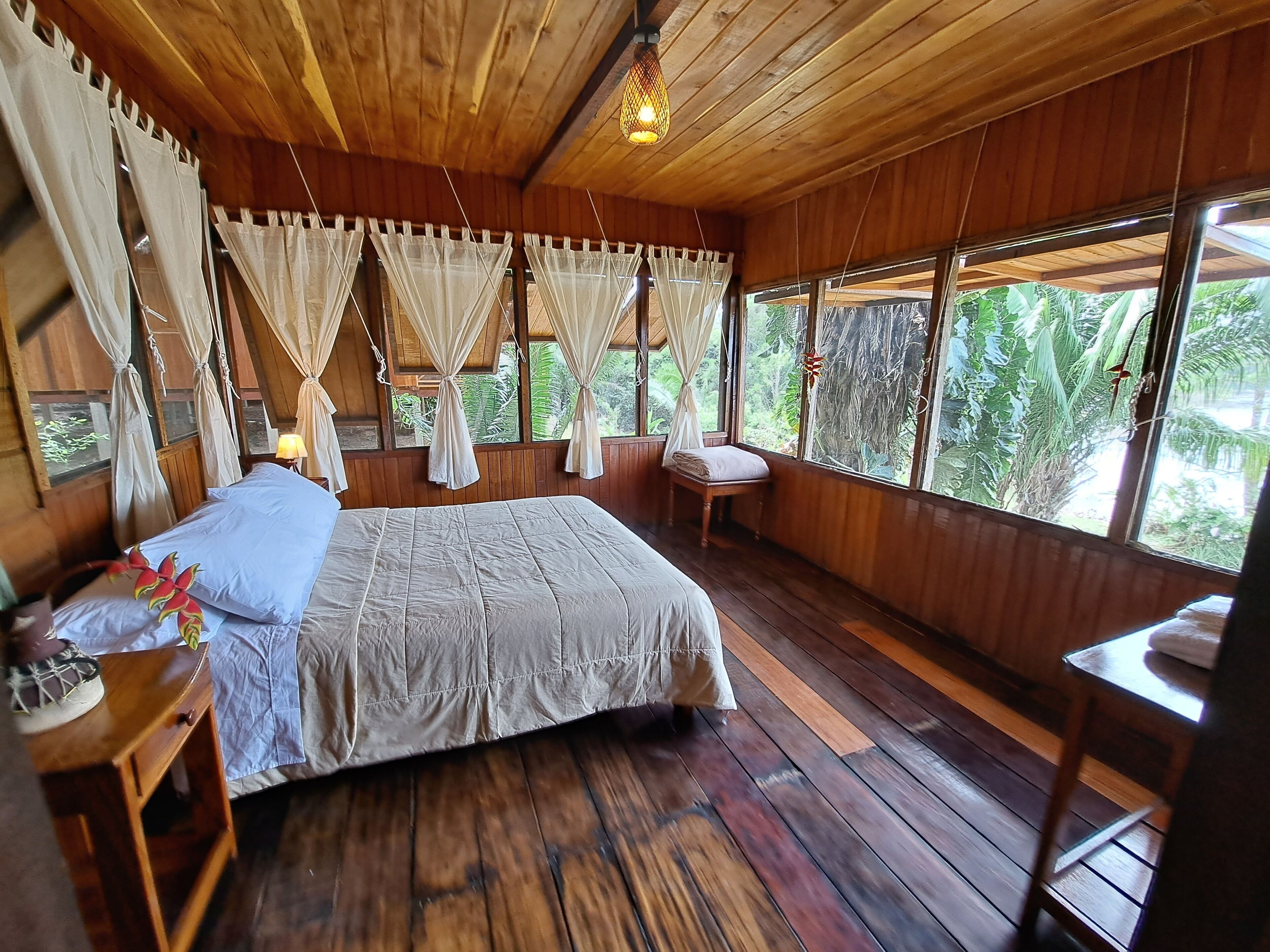 A wooden room with a bed, a nightstand, a chair, and a window with curtains.