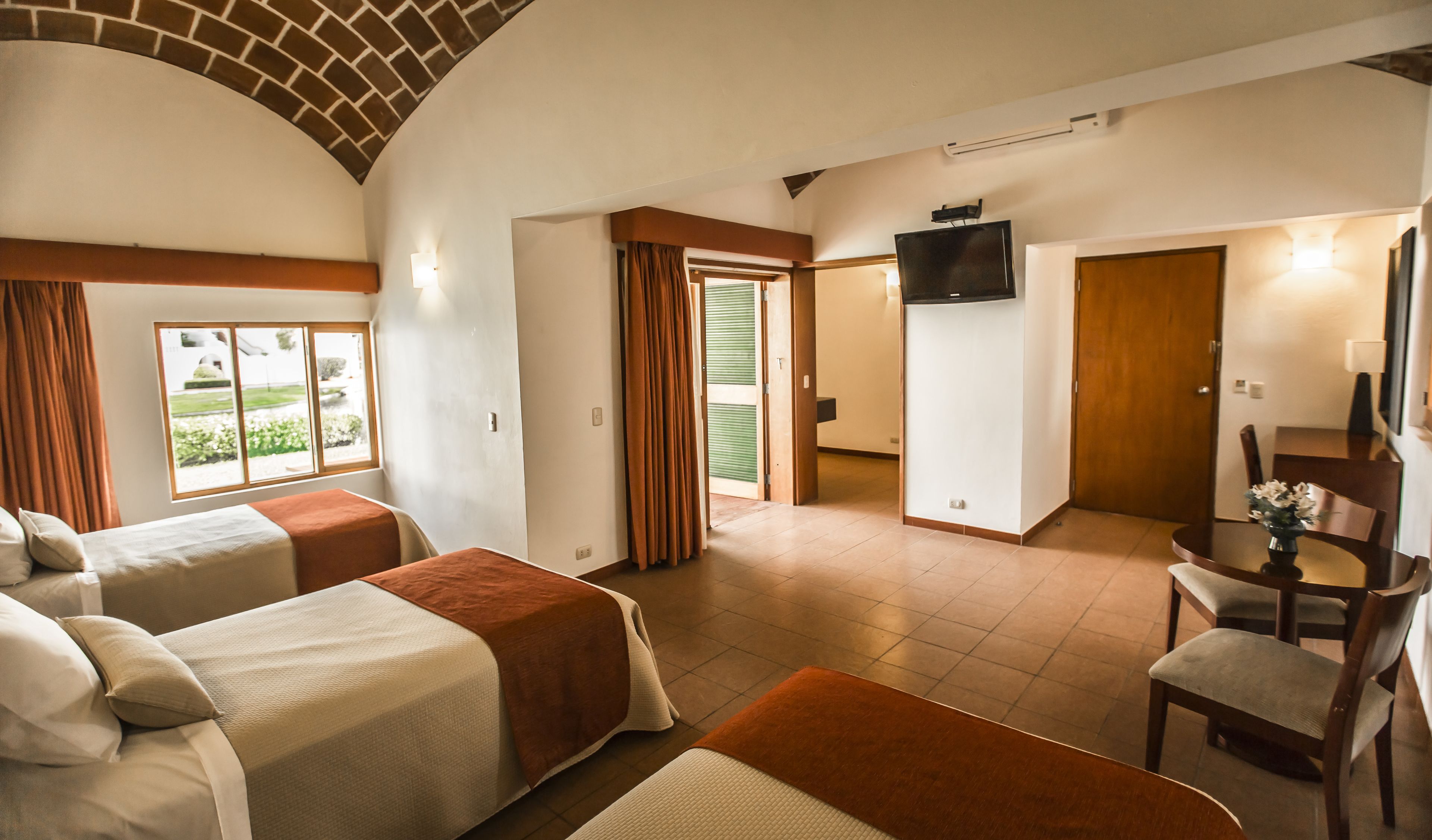 A hotel room with two beds, a TV, a dining area, and a view of the outdoors.