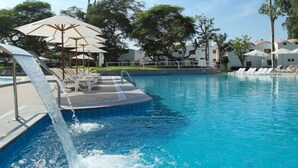 A swimming pool with a water feature, lounge chairs, umbrellas, and a building in the background. - Hotel Las Dunas (Ica)