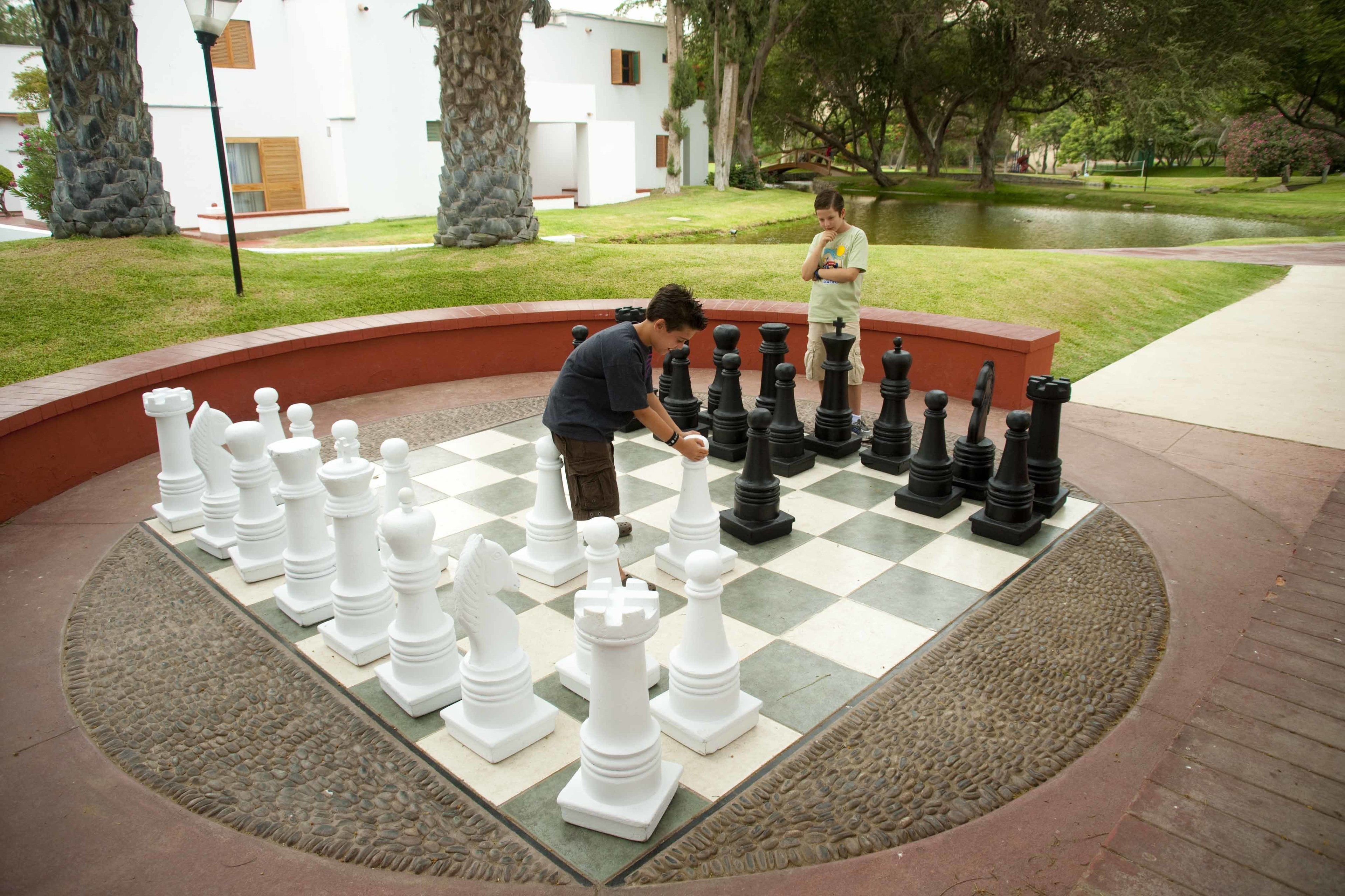 Un grand échiquier installé en plein air, avec des pièces d’échecs de taille surdimensionnée, entouré d’un espace paysager agrémenté d’arbres et d’un bassin.