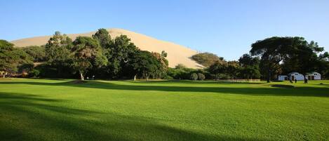 A golf course with a sand dune in the background, surrounded by trees and a few buildings.