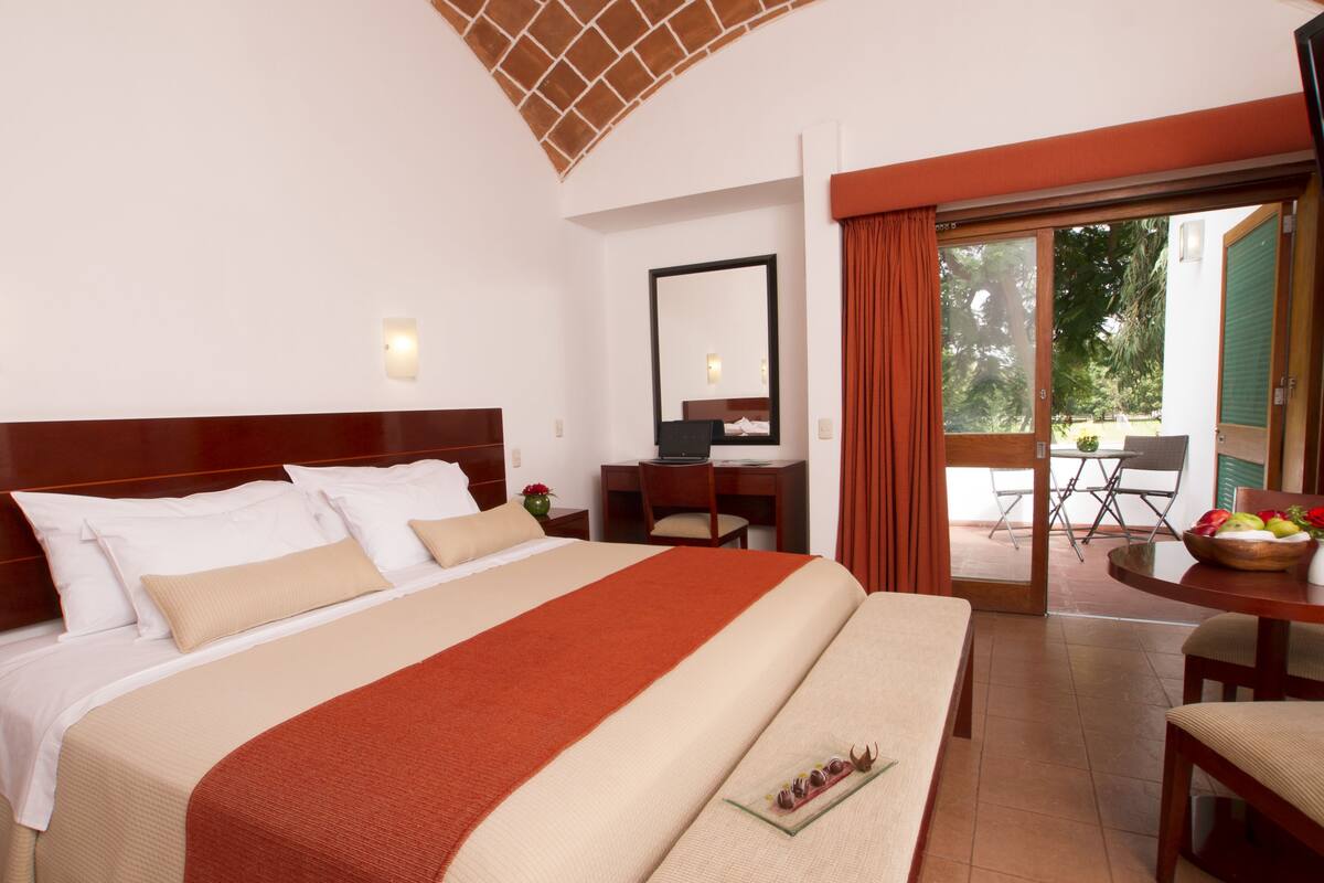 A hotel room with a large bed, a desk, a chair, and a dining area with a table and chairs.
