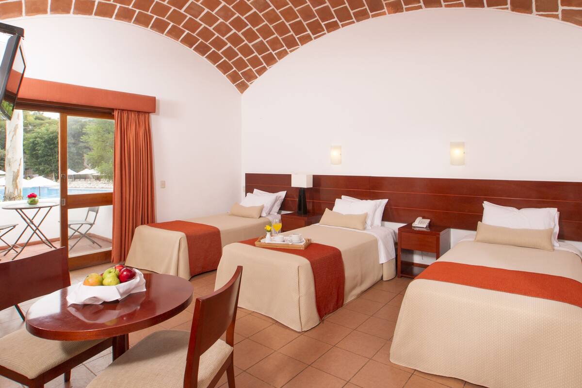 A hotel room with two single beds, a dining table with fruit, and a view of the outdoors.