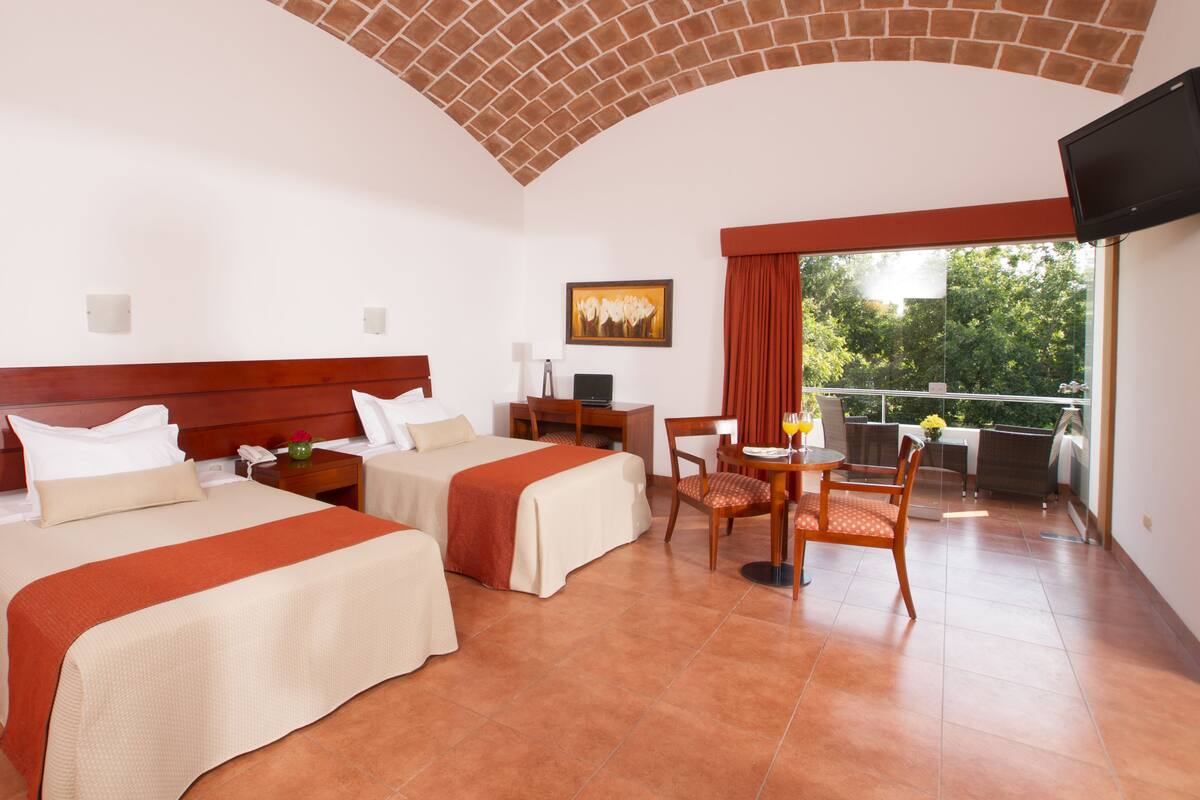 A hotel room with two beds, a desk, a chair, a TV, and a balcony with a view of trees.