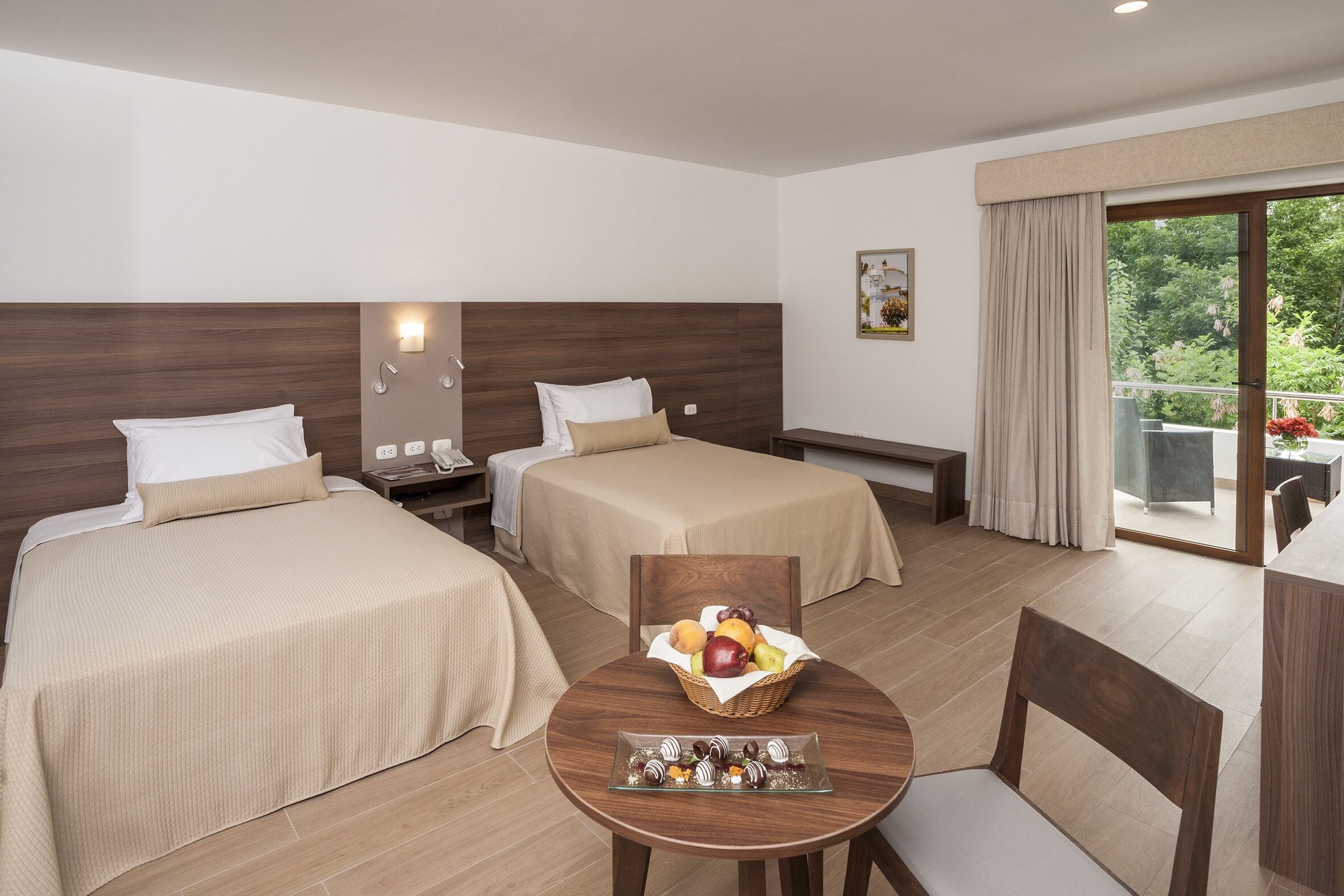 Habitación de hotel con dos camas, una mesa de comedor y una cesta de fruta, y vistas a la vegetación por la ventana.