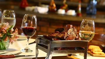 Una mesa de comedor con una bandeja metálica que sostiene un filete, dos copas de vino, un jarrón con flores y un plato de pan.