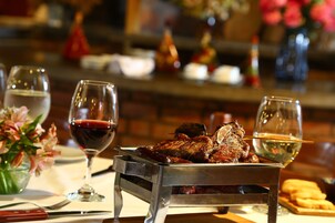 Una mesa de comedor con una bandeja metálica que sostiene un filete, dos copas de vino, un jarrón con flores y un plato de pan.