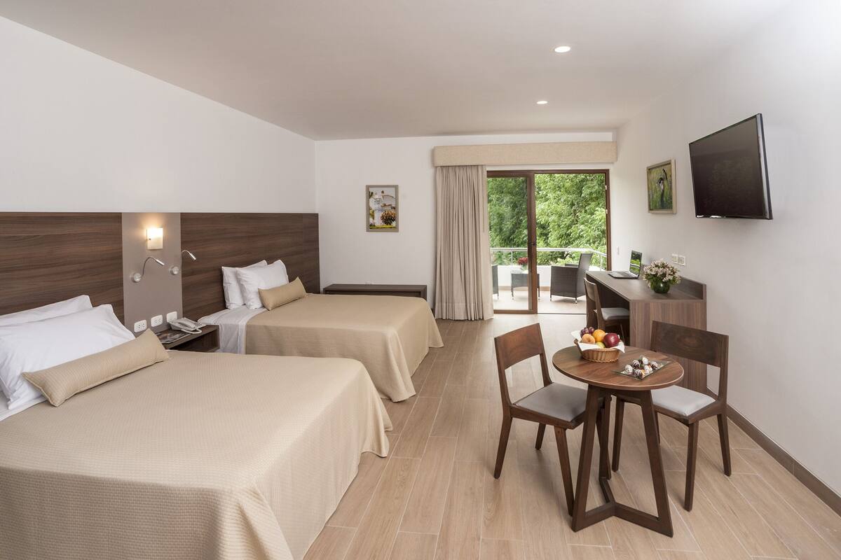 A hotel room with two beds, a dining table with chairs, a TV, and a balcony with a view of greenery.