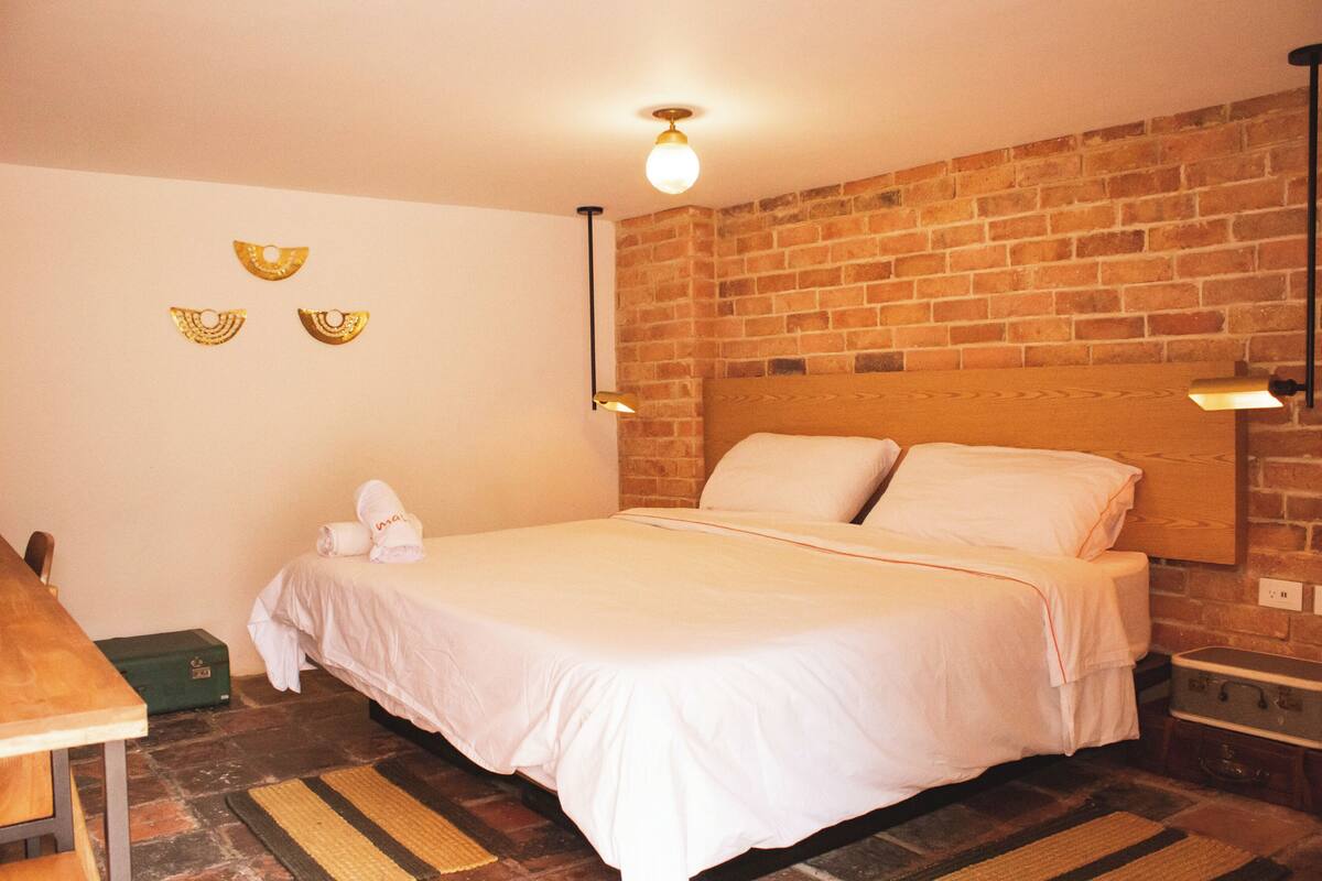 A hotel room with a brick wall, a bed with white bedding, a wooden headboard, a small table, and a green suitcase.