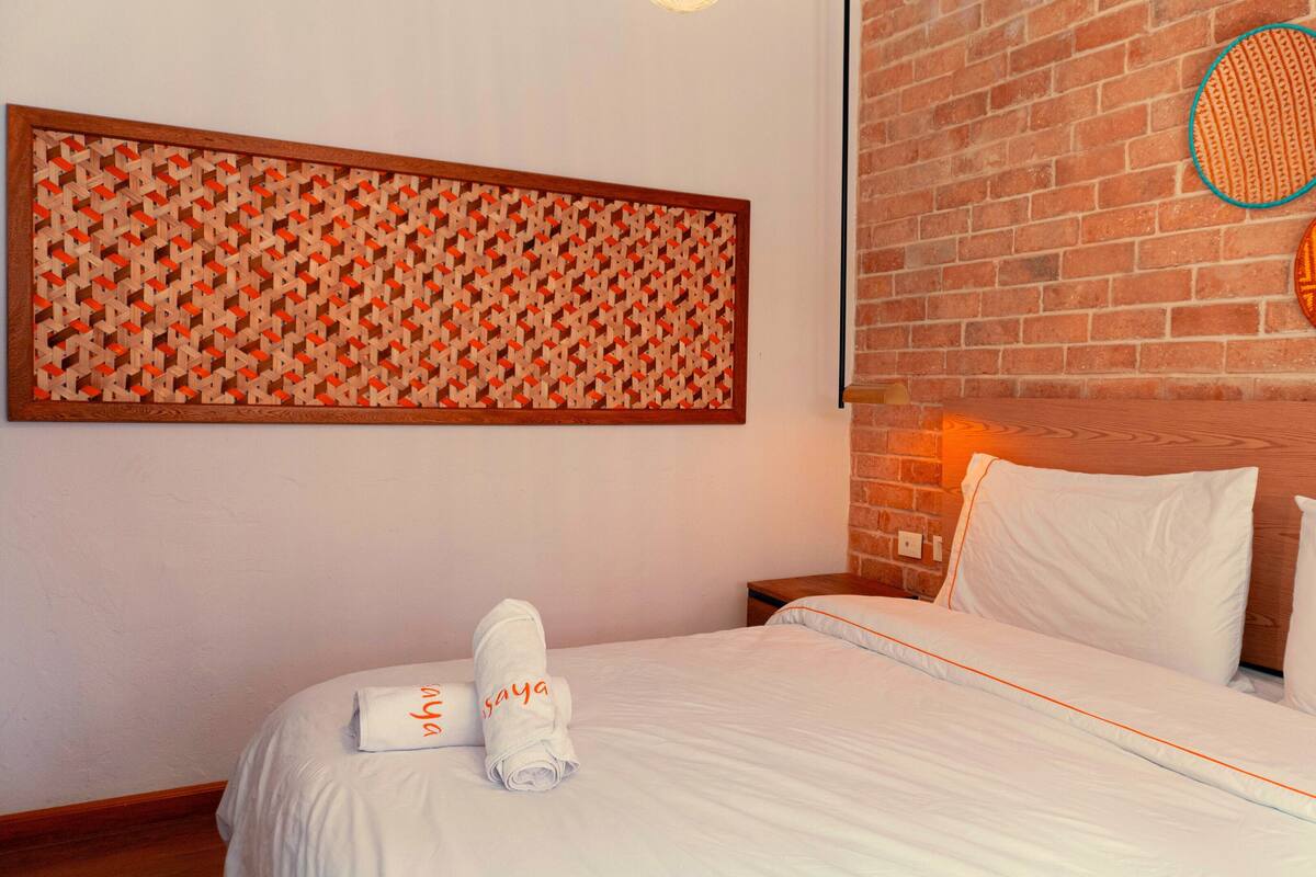 A neatly made bed with white linens and two towels with text, a patterned wall hanging, and a brick wall with decorative plates.