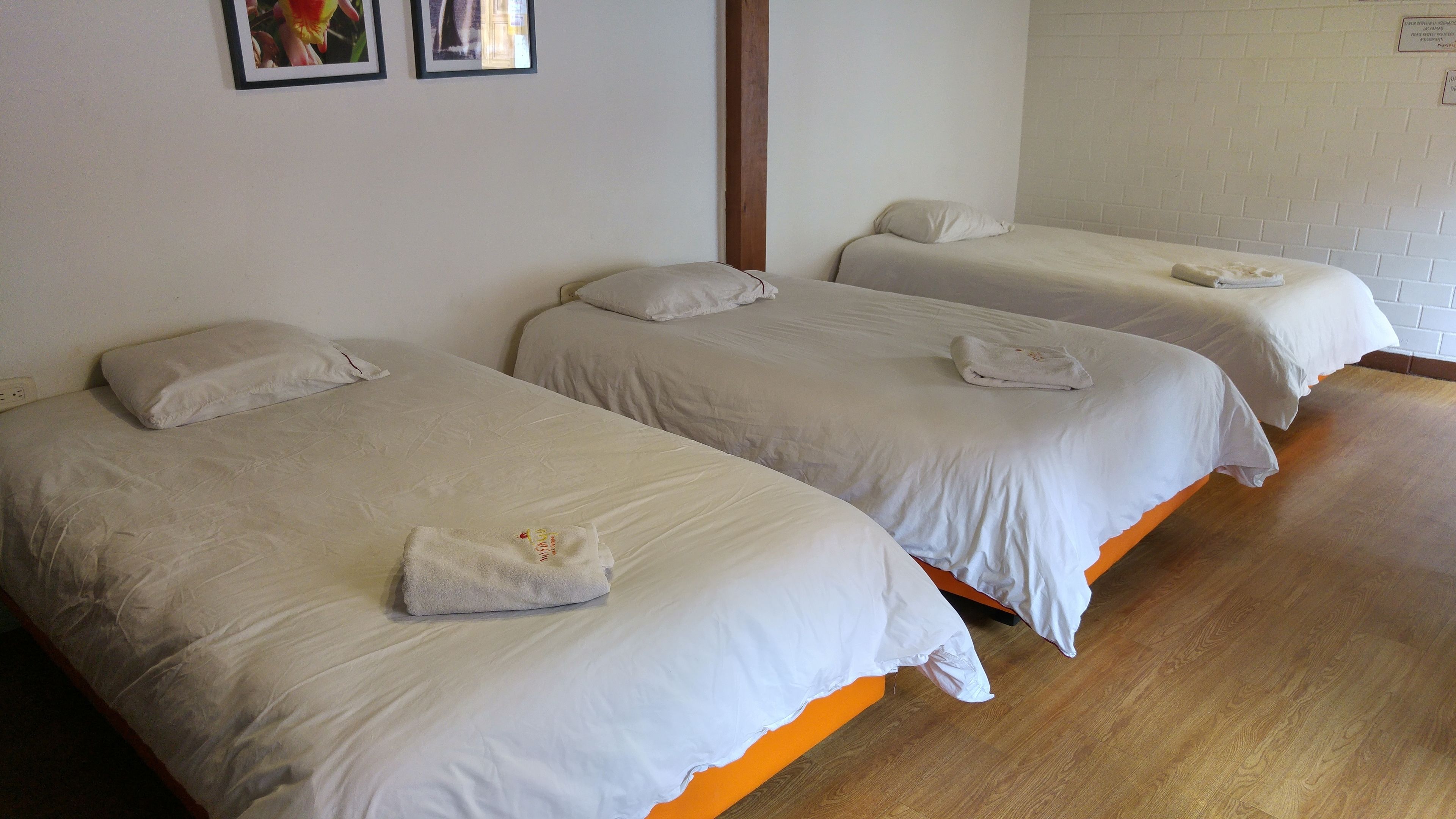 Trois lits simples avec du linge de lit et des serviettes blancs, un plancher en bois et un mur en briques blanches.