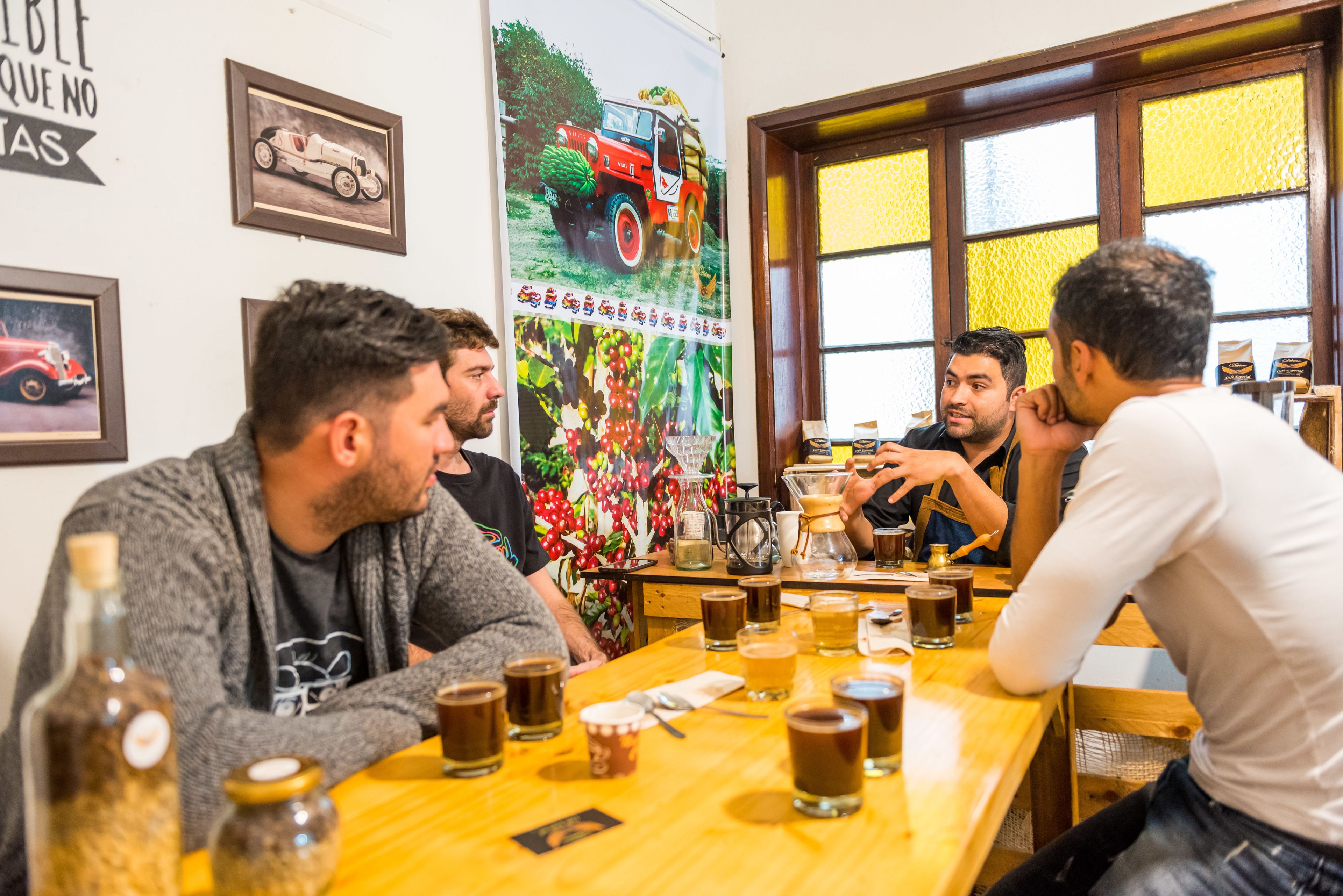 A group of people sitting around a wooden table with various drinks and a coffee pot, in a room with framed pictures on the wall and a window with yellow curtains.