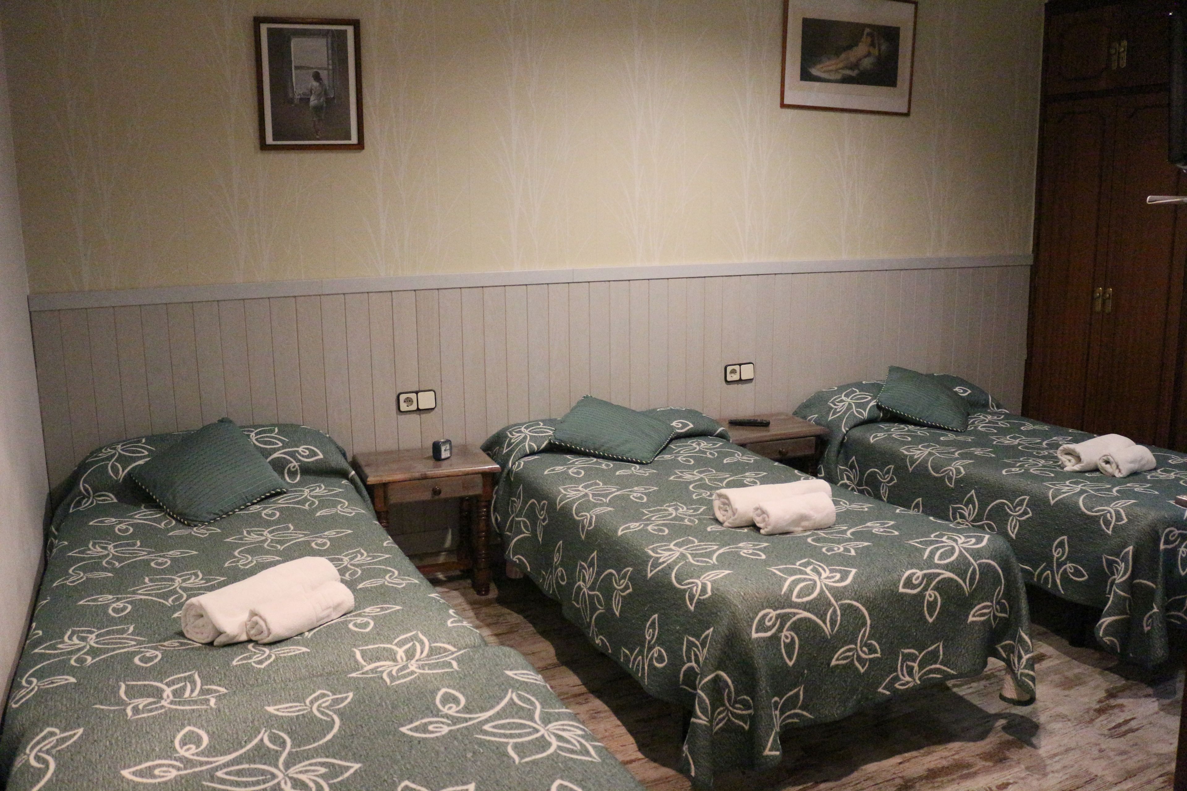 A hotel room with three single beds, each with a green floral bedspread and a white towel. There are two bedside tables with lamps, and the walls are adorned with framed pictures.