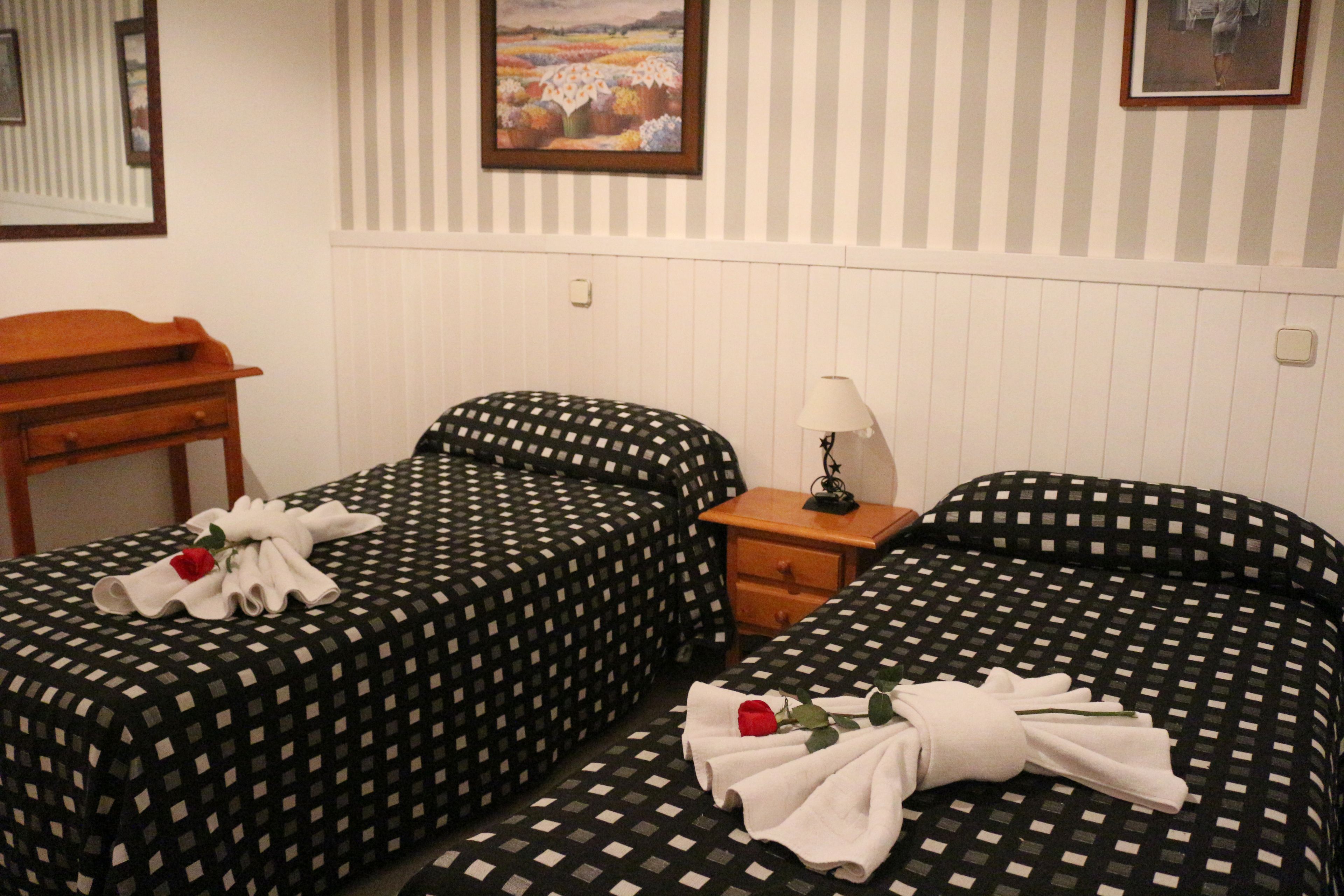 A room with two beds, each with a checkered bedspread and a single white towel with a red rose. There is a wooden nightstand with a lamp between the beds.