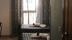 Habitación de hotel con dos camas, una ventana con cortinas estampadas, una puerta de madera y una pequeña mesa con una flor roja.