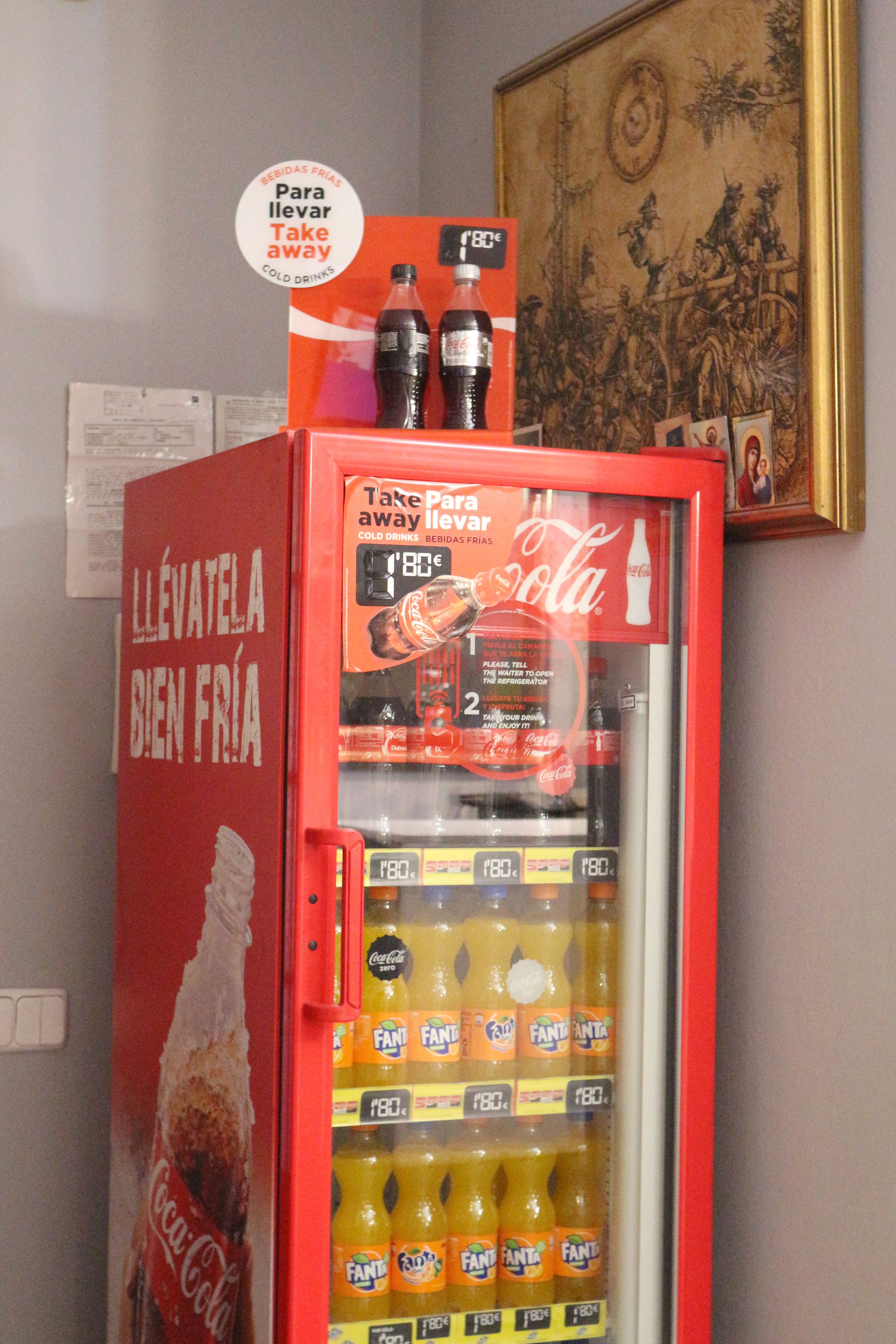 A red Coca-Cola vending machine with Fanta bottles inside, located in a room with a painting on the wall.