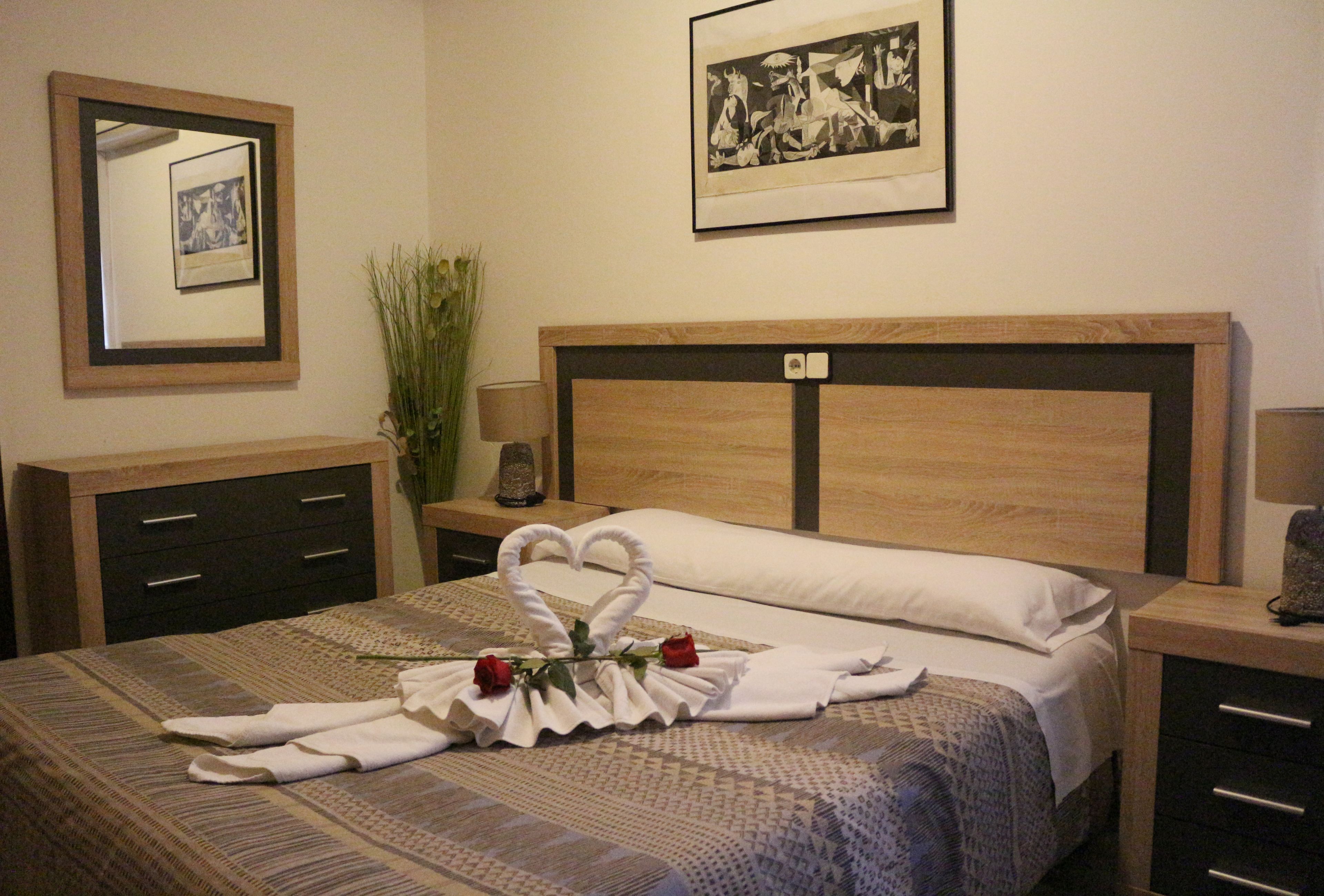 Ein ordentlich gemachtes Bett mit Herzchen-Handtuch-Arrangement und Rosen, eine Nachttischlampe, ein gerahmtes Bild an der Wand und eine kleine Pflanze.