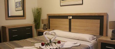 A neatly made bed with a heart-shaped towel arrangement and roses, a bedside lamp, a framed picture on the wall, and a small plant.