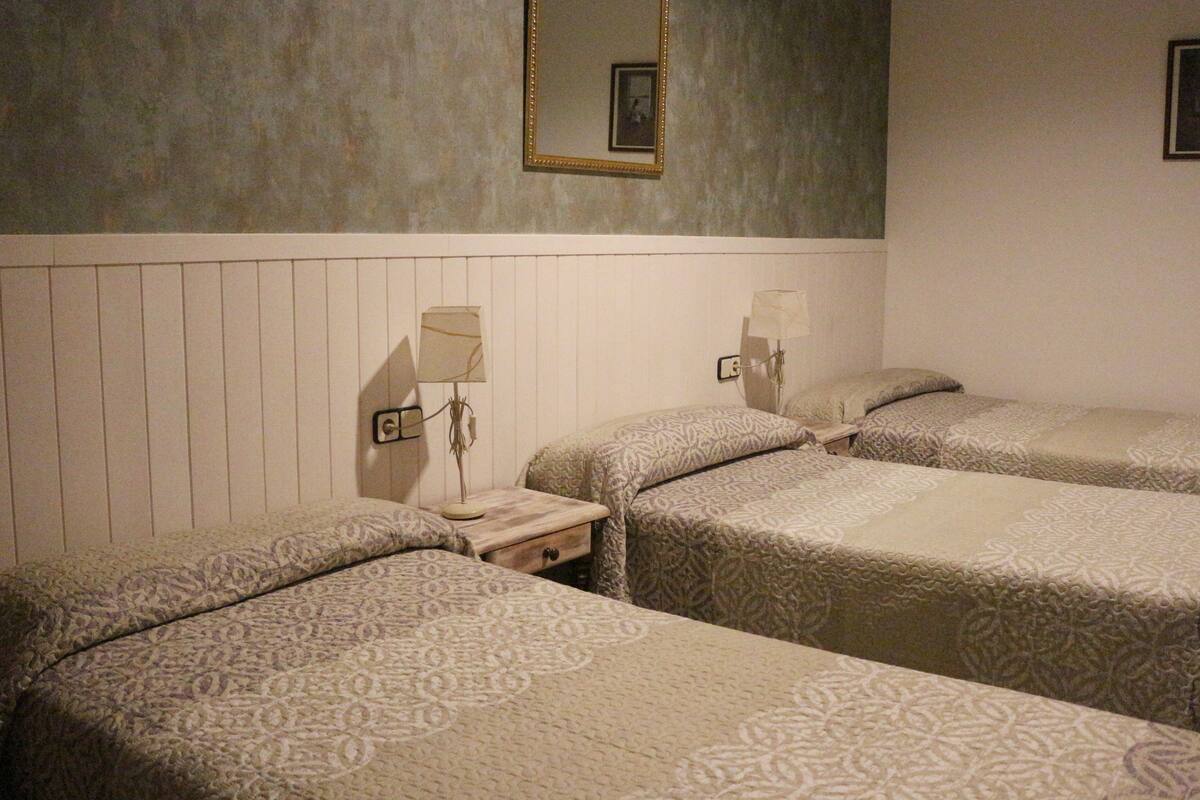 A hotel room with three single beds, a bedside table with a lamp, a wall-mounted mirror, and a framed picture.