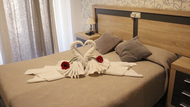 Una cama bien hecha con cabecera estampada con motivos florales, una mesita de noche con lámpara y un arreglo decorativo de toallas con flores.