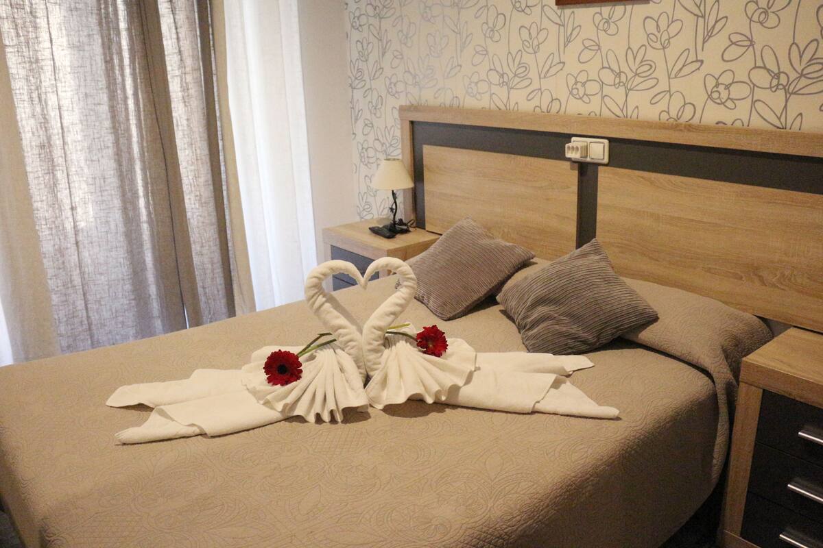 A neatly made bed with a floral-patterned headboard, a nightstand with a lamp, and a decorative towel arrangement with flowers.