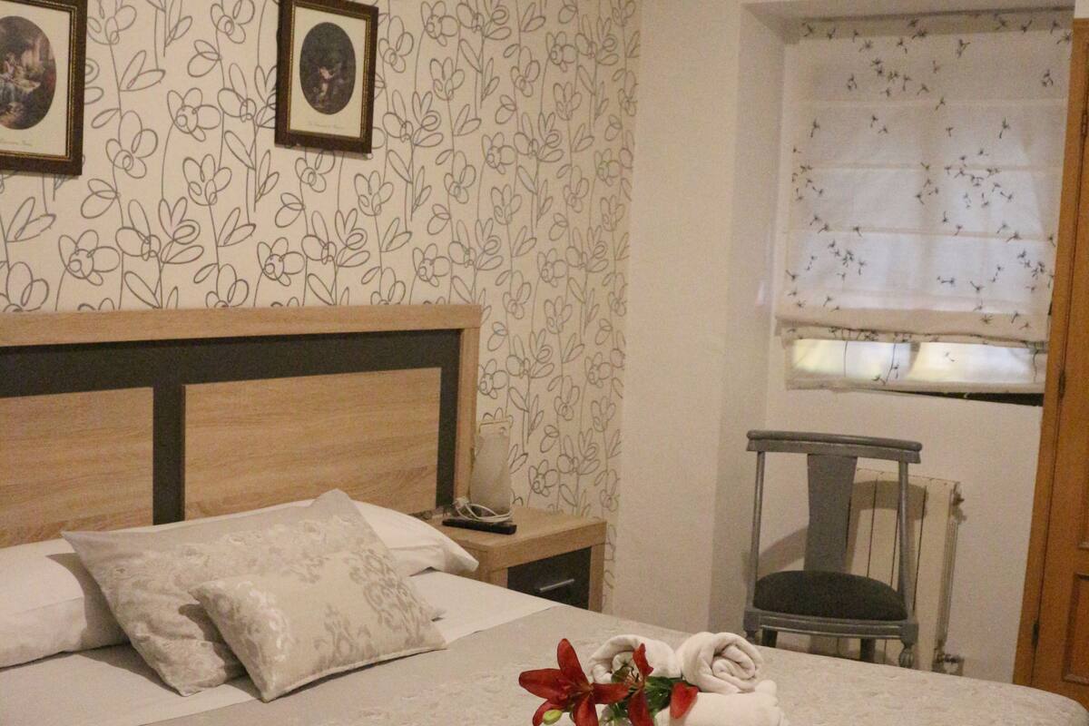 A neatly made bed with a floral-patterned headboard, a small table with a chair, and a window with a view of birds.