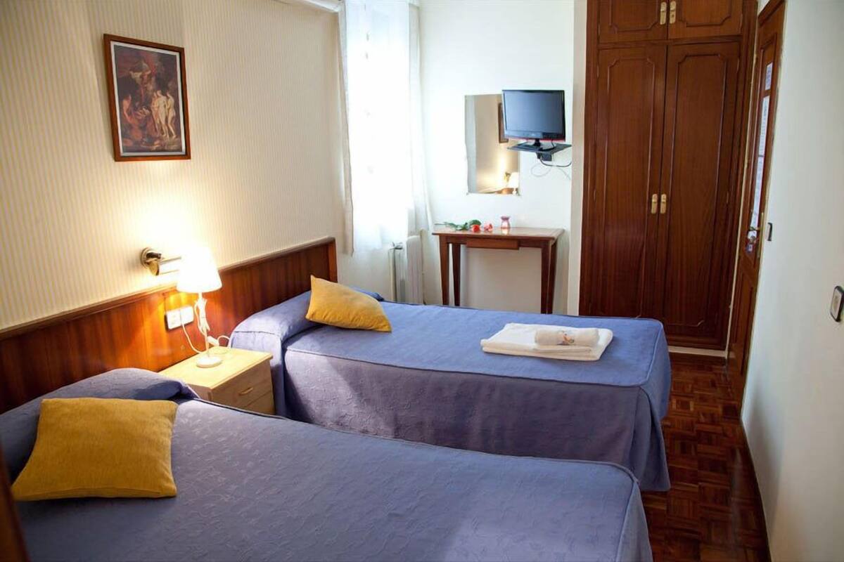 A hotel room with two beds, a bedside table with a lamp, a small table with a TV, and a wooden wardrobe.
