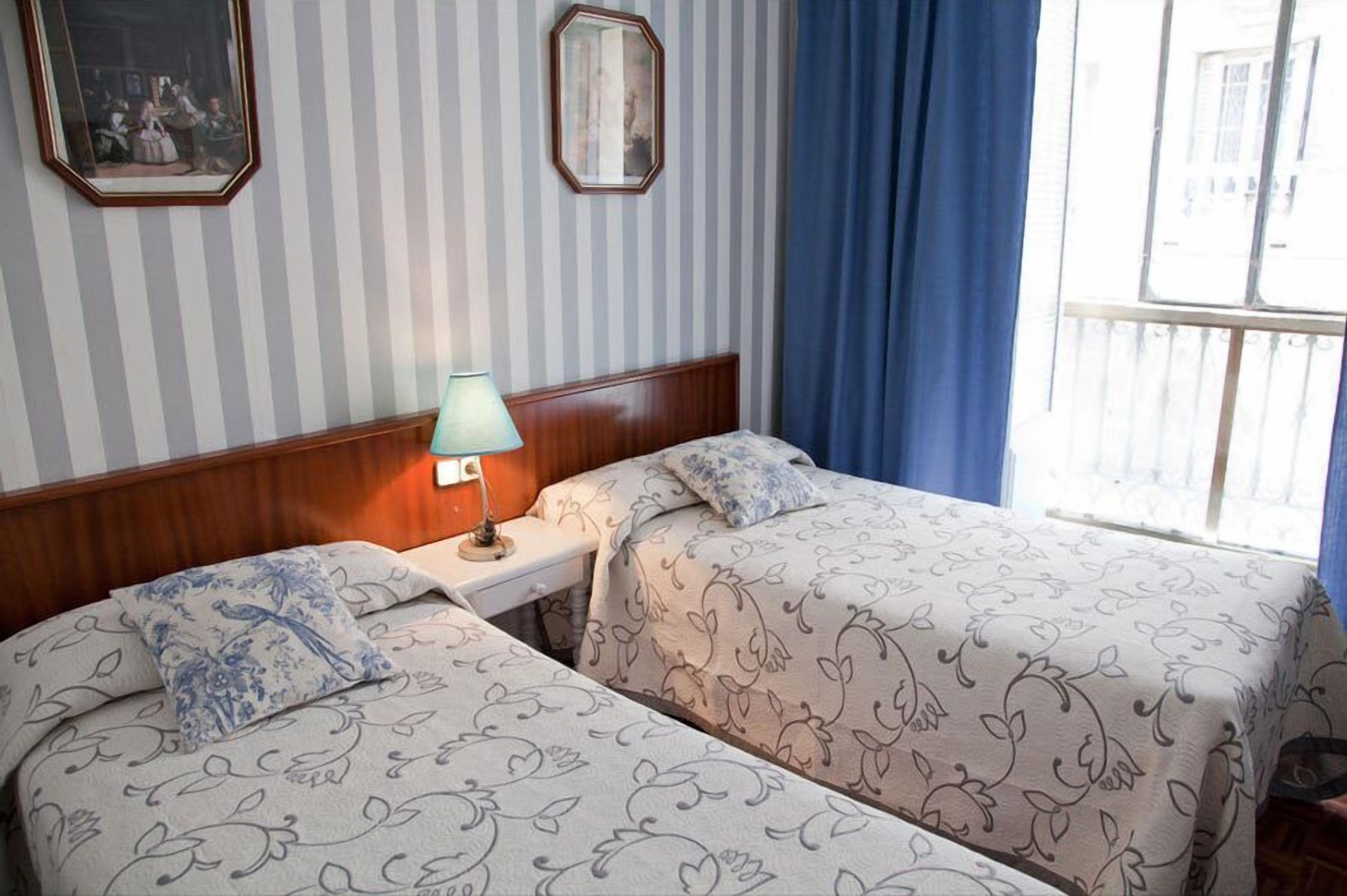 A hotel room with two beds, striped wallpaper, a wooden headboard, a nightstand with a lamp, and a window with blue curtains.
