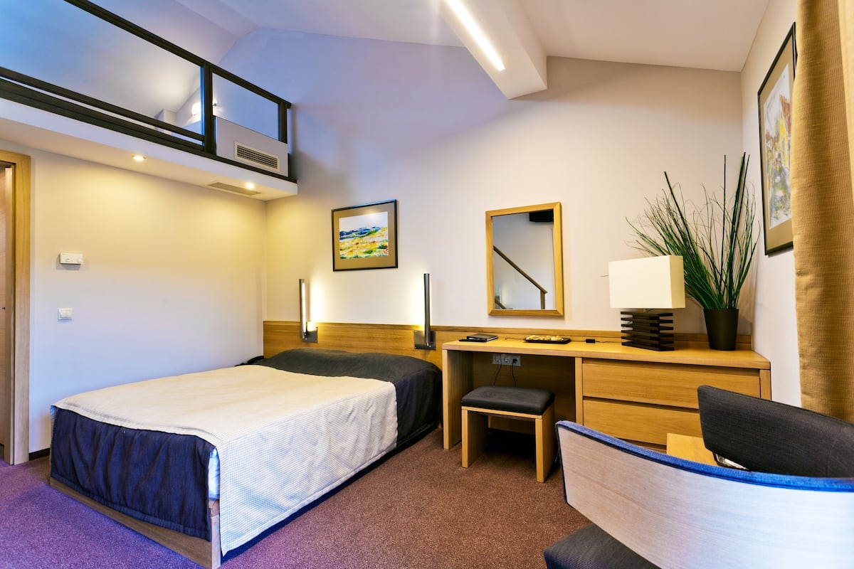 Habitación de hotel con cama, escritorio, silla y una mesita de noche con lámpara.