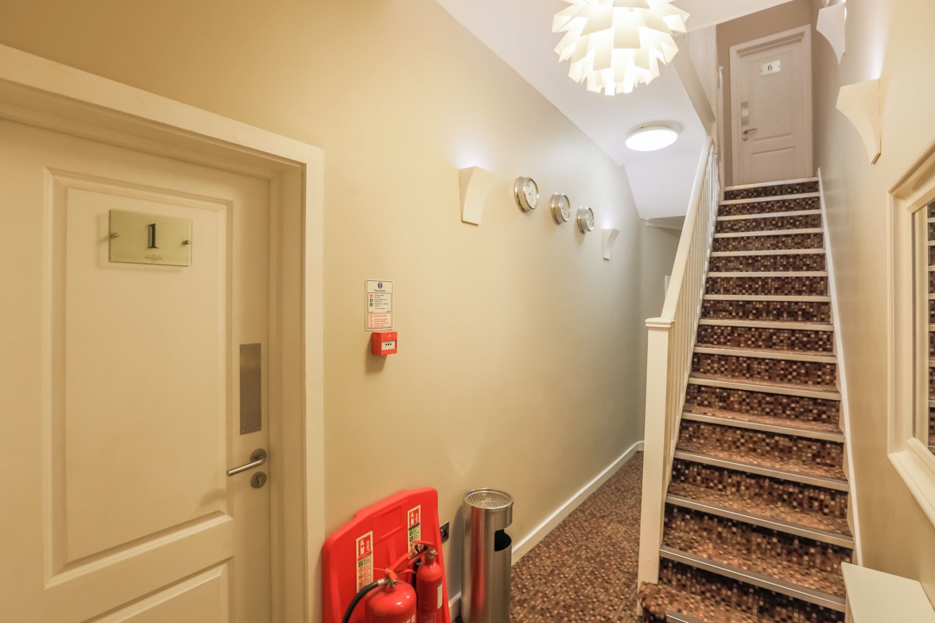A hallway with a door marked '1', a fire extinguisher, a trash can, and a staircase with a patterned carpet.