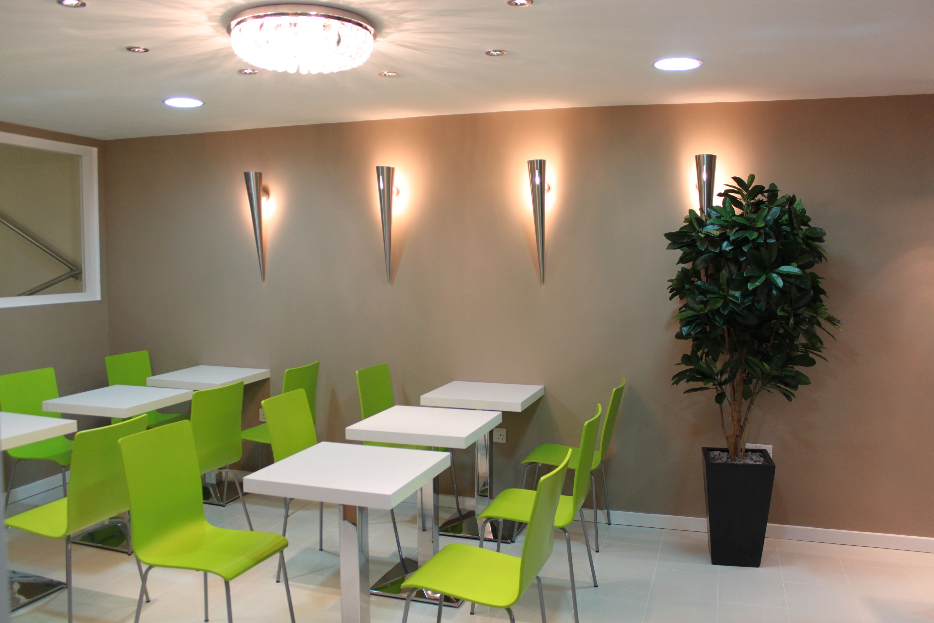 A room with green chairs and white tables, a potted plant, and wall-mounted lights.
