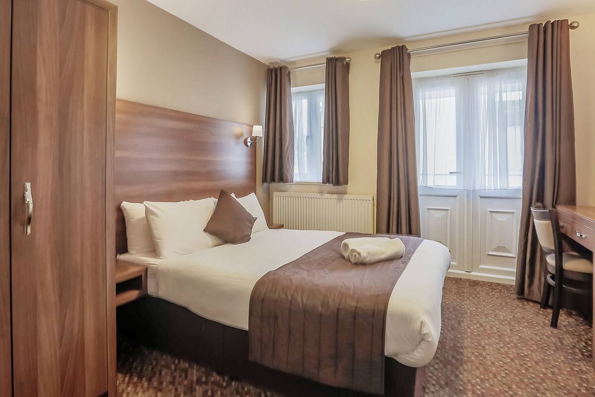 Ett hotellrum med en stor säng, en sänggavel i trä, ett skrivbord och ett fönster med gardiner.