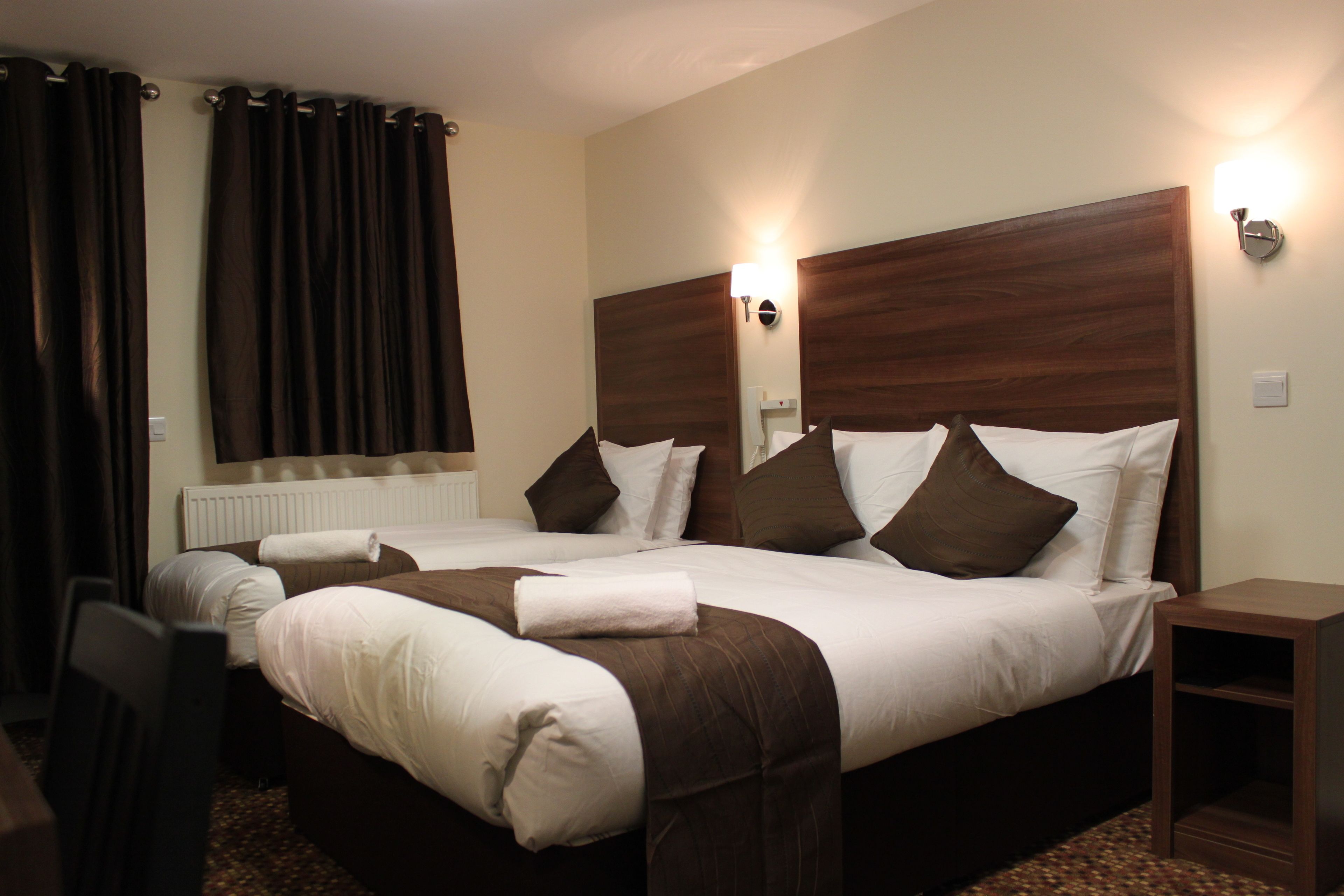 A hotel room with two beds, dark curtains, a wooden headboard, a nightstand, and a wall-mounted light fixture.