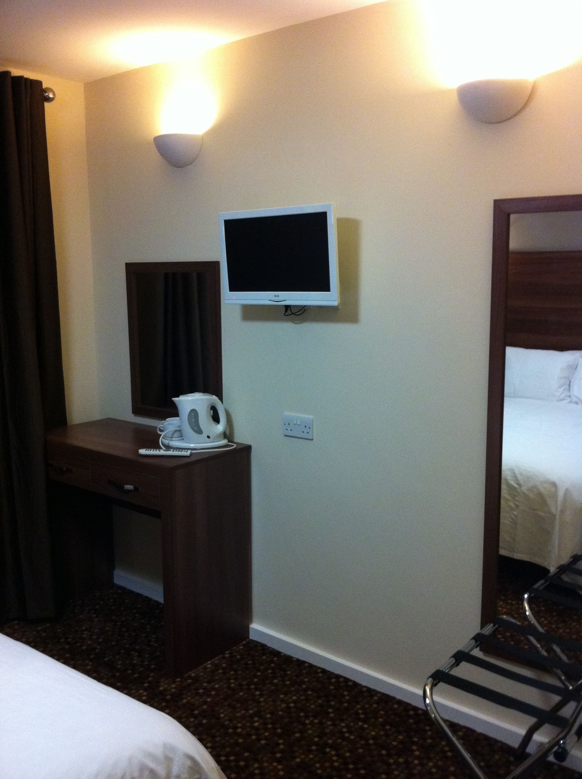 A hotel room with a bed, a desk with a telephone and a kettle, a television mounted on the wall, and a mirror.