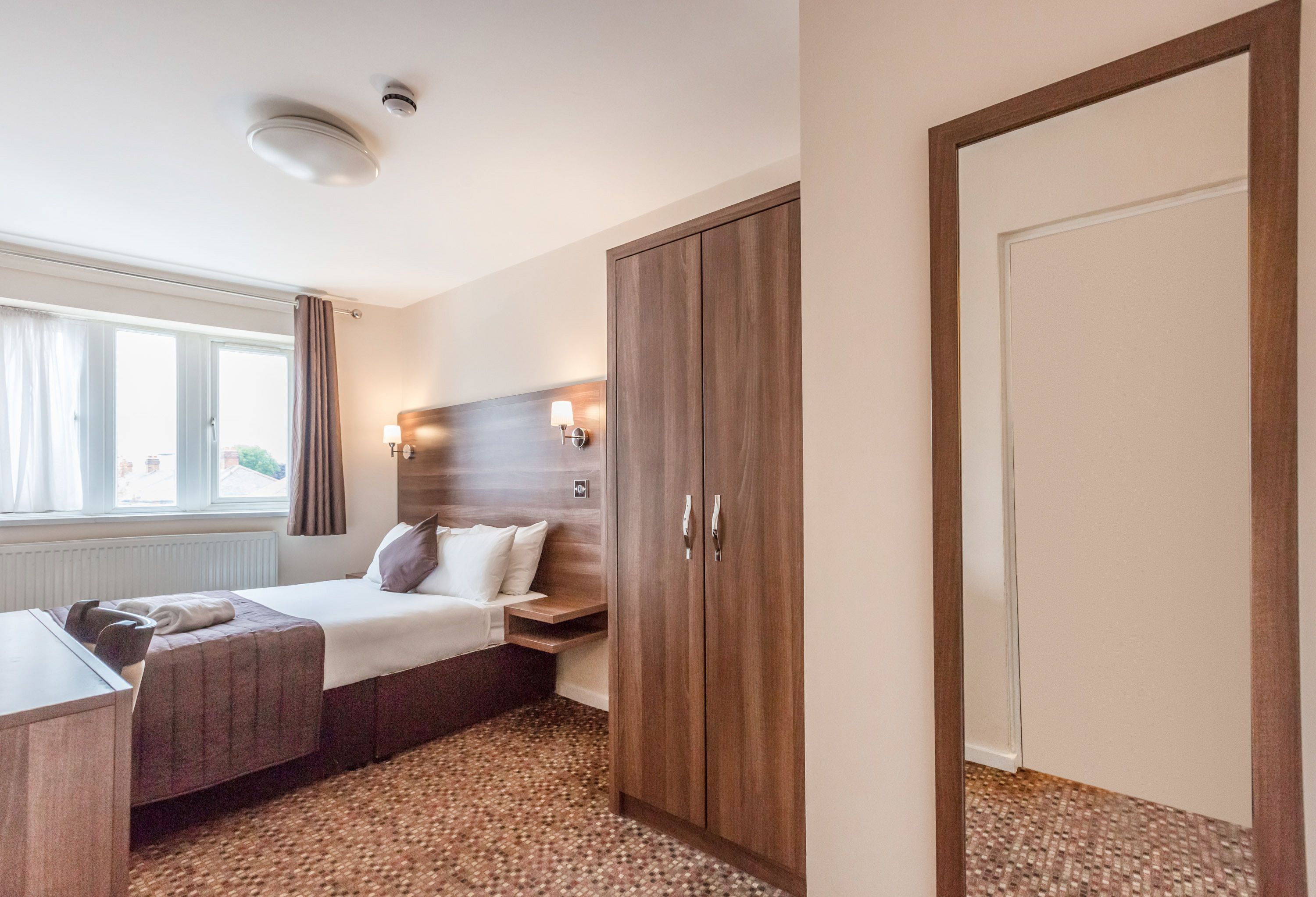 A hotel room with a bed, bedside table, lamp, wardrobe, and a window with curtains.