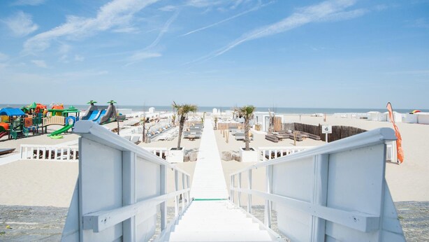 A beachside walkway leading to a sandy beach with a playground and palm trees. - Beach Hotel Helios by CW Hotel Collection (Blankenberge)