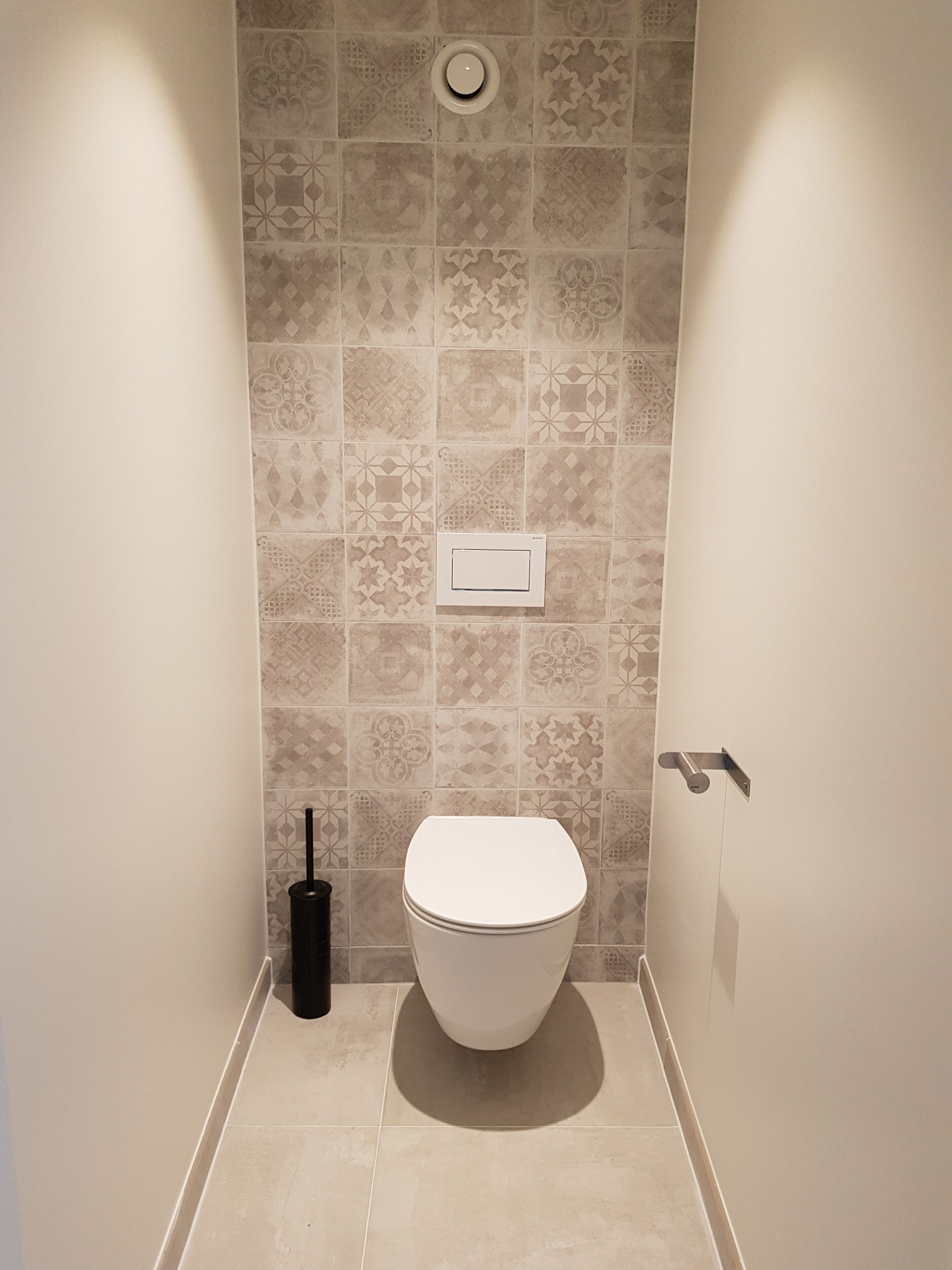 A bathroom with patterned tiles, a toilet, a hand dryer, and a wall-mounted flush handle.