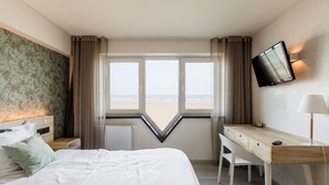 A hotel room with a bed, a desk, a chair, a TV mounted on the wall, and a view of the beach through large windows. - Beach Hotel Helios by CW Hotel Collection (Blankenberge)