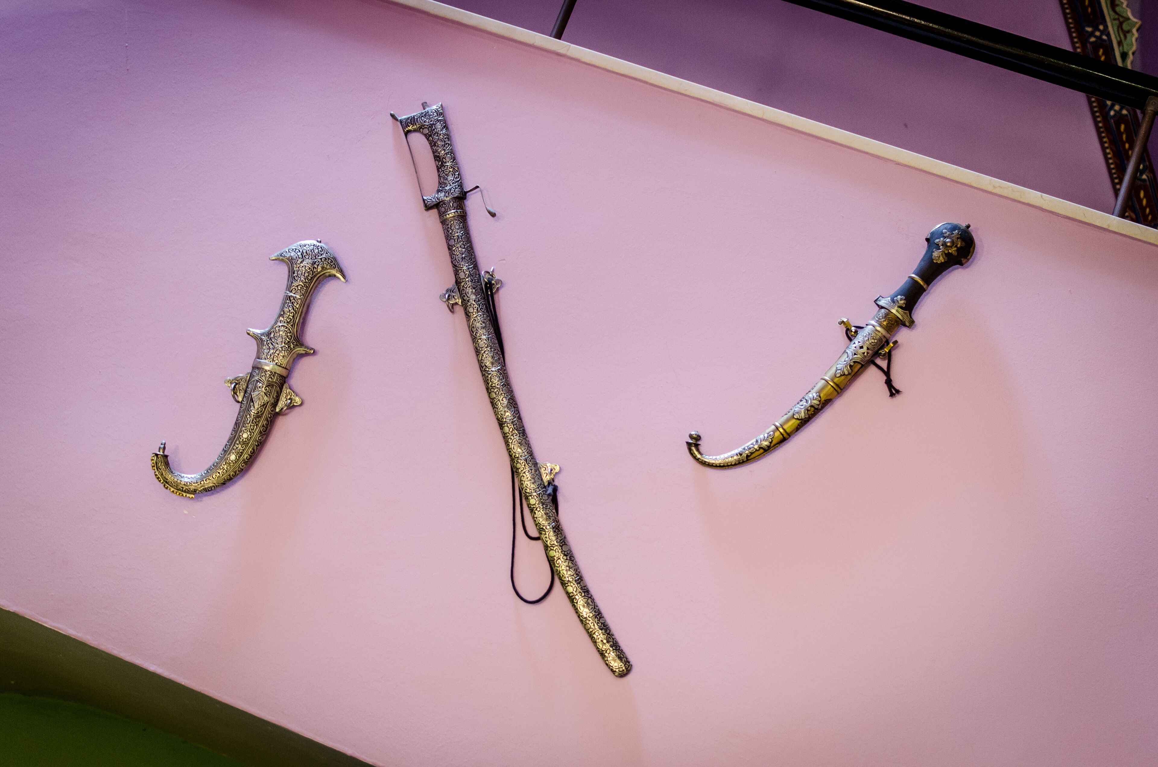 Three ornate swords displayed on a pink wall.