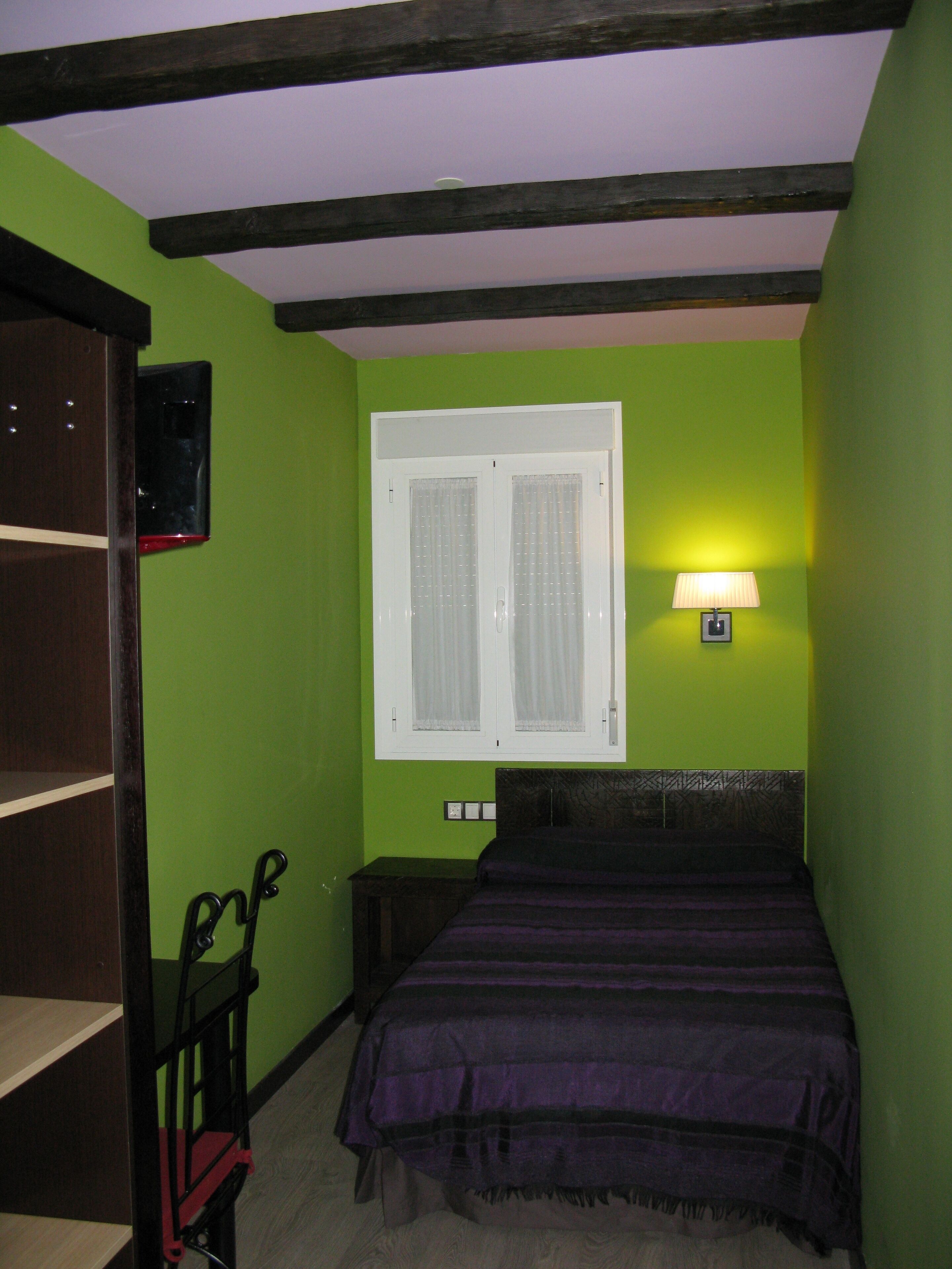 A bedroom with a bed, a window with curtains, a nightstand, a lamp, and a shelf with a mirror.