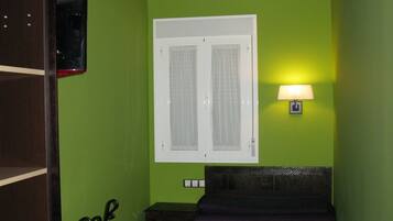 A bedroom with a bed, a window with curtains, a nightstand, a lamp, and a shelf with a mirror.
