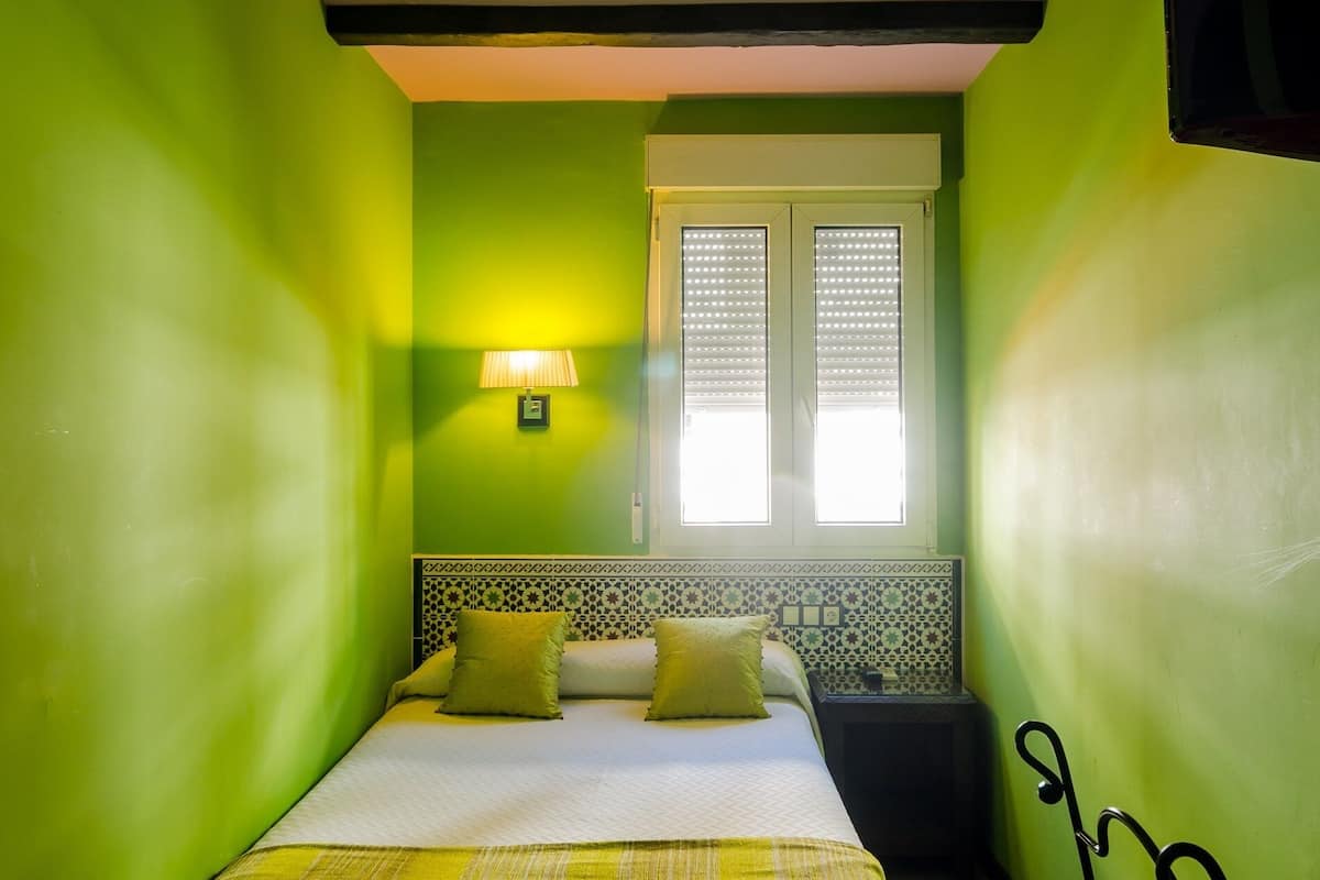 A room with green walls, a bed with a yellow bedspread, a nightstand, a lamp, and a window with blinds.