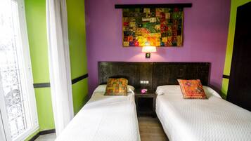 A room with two beds, a colorful quilted wall hanging, and a window with curtains.