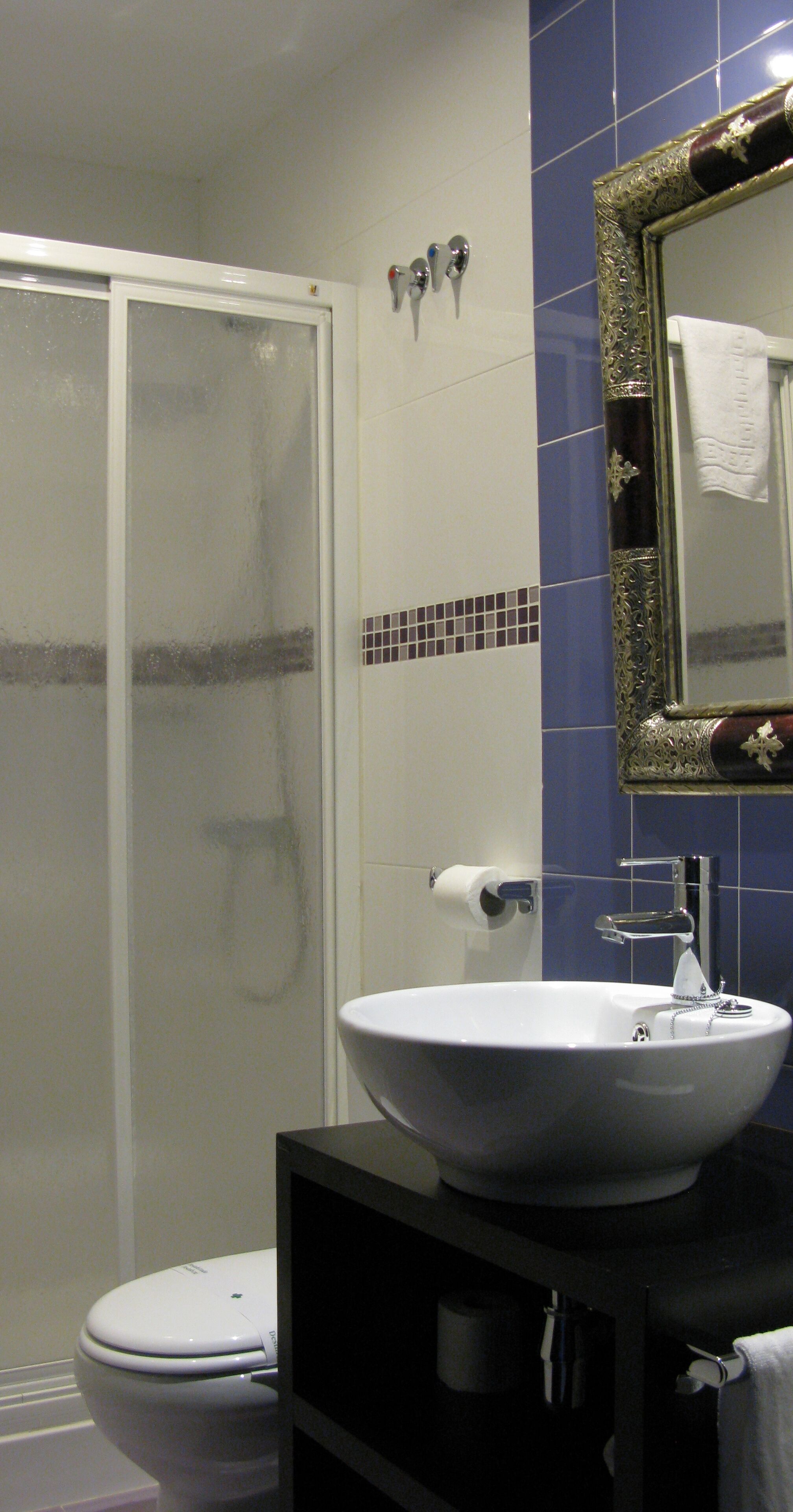 Un baño con inodoro blanco, un lavamanos blanco con encimera negra, cabina de ducha de vidrio y un espejo con marco decorativo.
